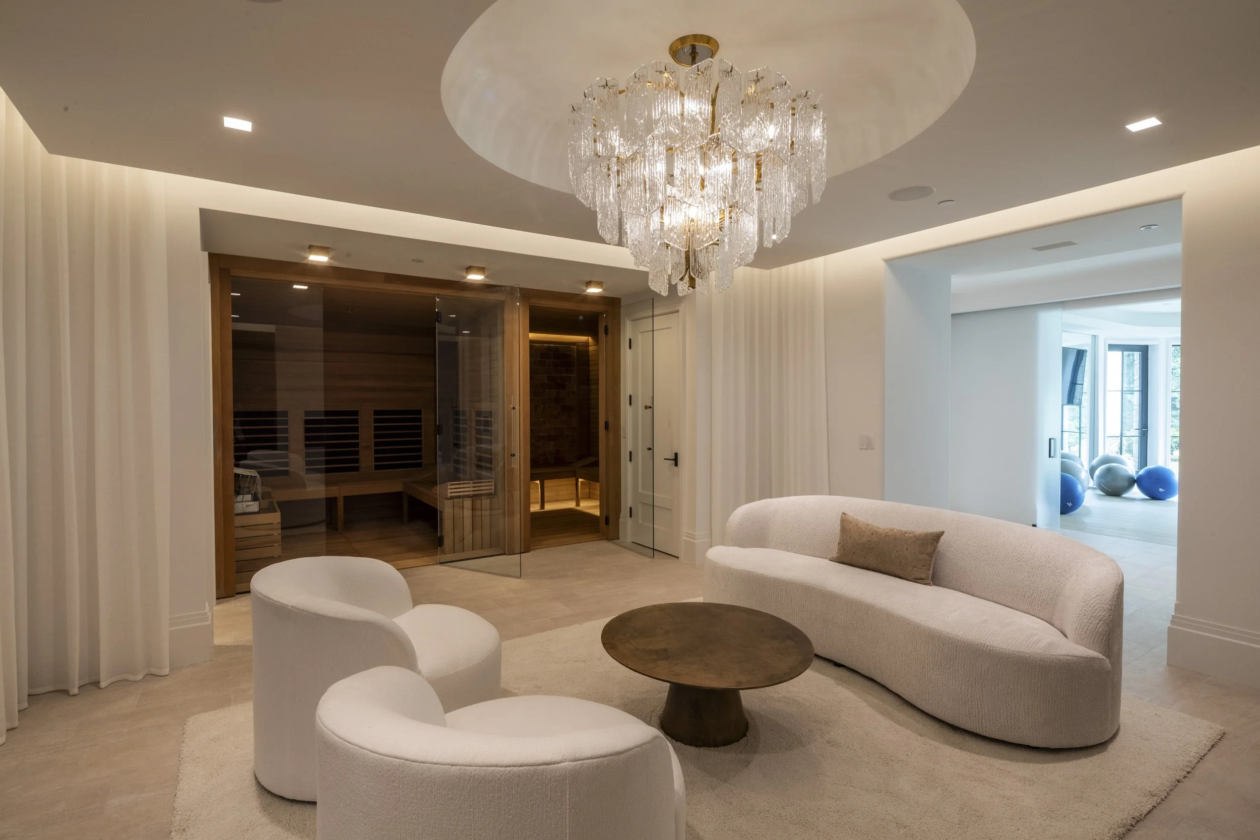 Living room with a chandelier above a beige curved sofa and white chairs, a round wooden coffee table, a beige rug, and a ceiling with ambient lighting. In the background, there is a wooden sauna with a glass door and a fitness area with blue and whi