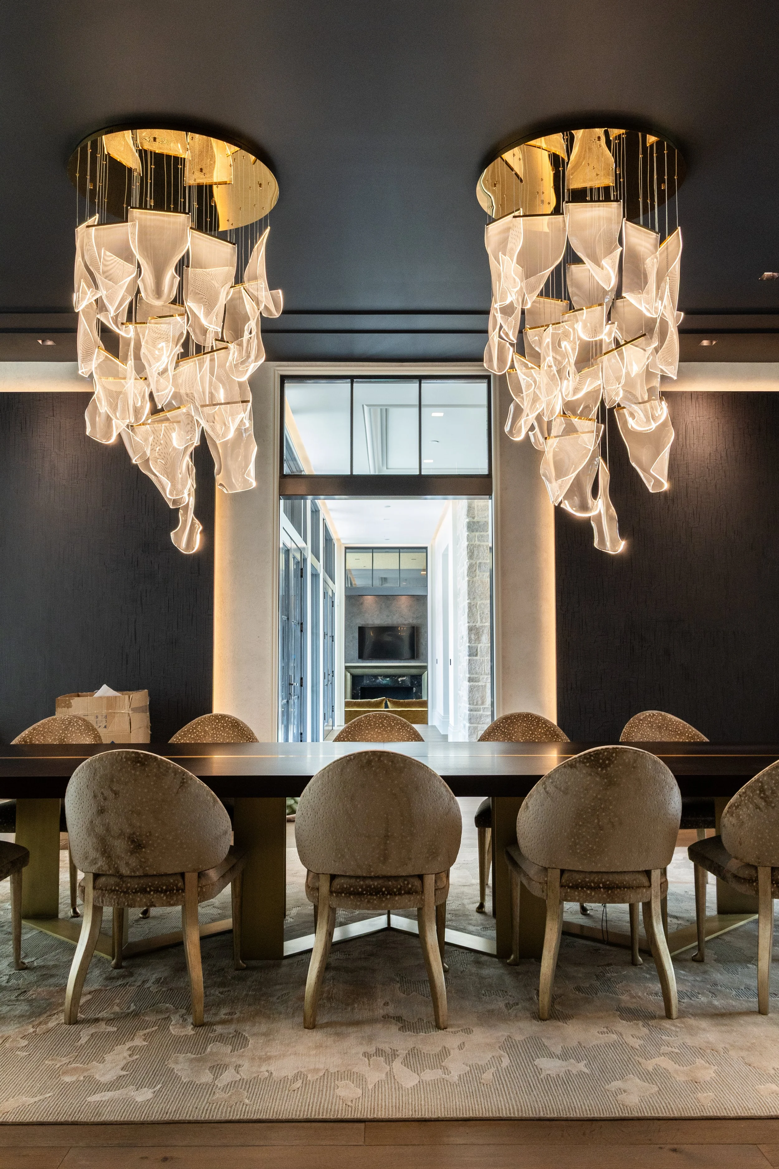 A modern chandelier with multiple white fabric or glass elements hanging from the ceiling over a large wooden dining table surrounded by upholstered chairs. There's a large window or mirror in the background reflecting the space, and the room has dar