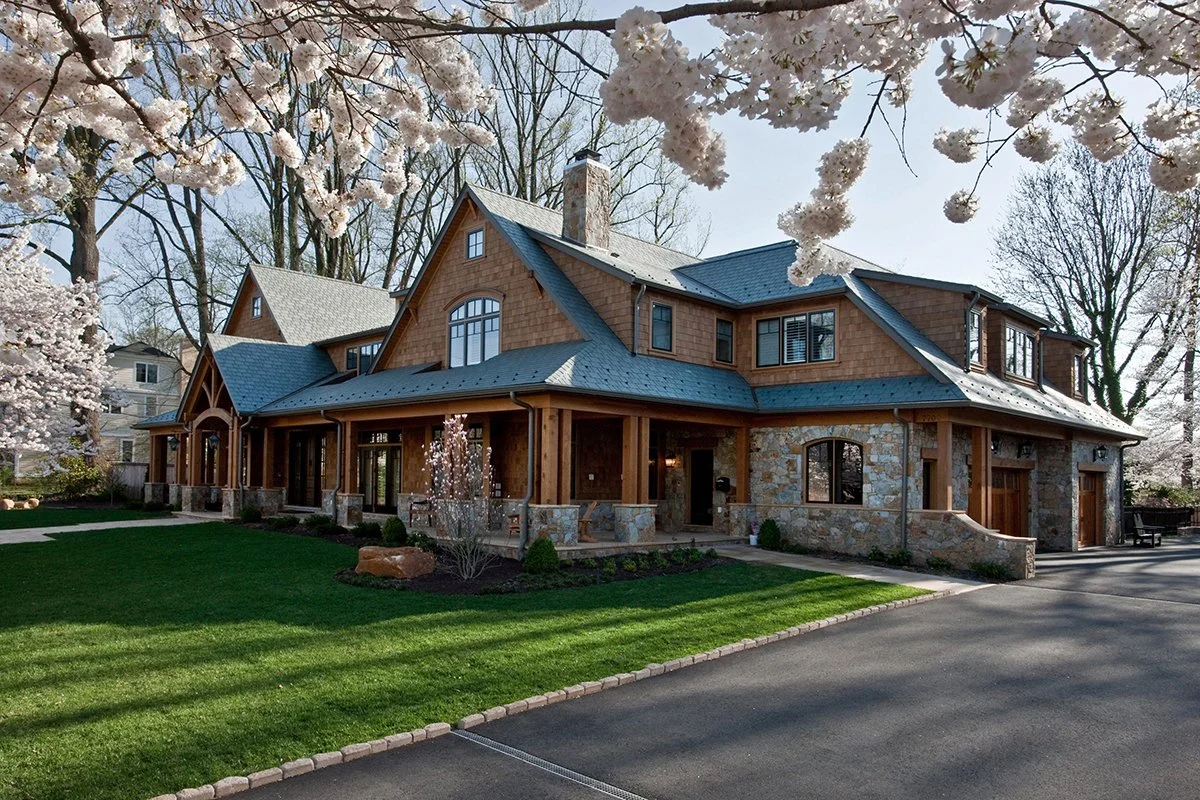 A large house with a mix of stone and wood exterior, surrounded by a well-maintained lawn and trees with blooming white flowers.