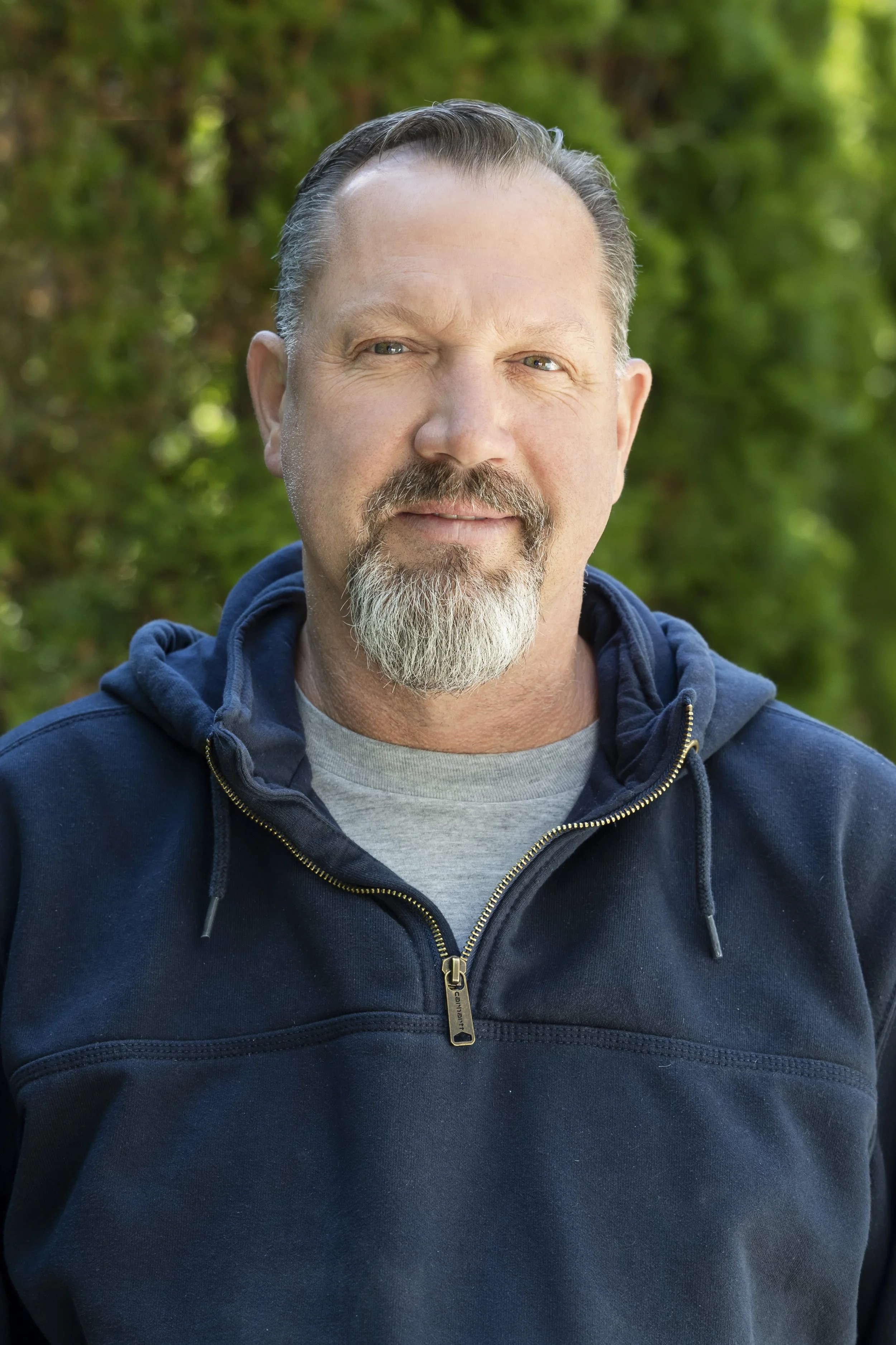 Close-up of a middle-aged man with gray hair and a beard, wearing a navy blue zip-up hoodie, standing outdoors with green trees in the background.