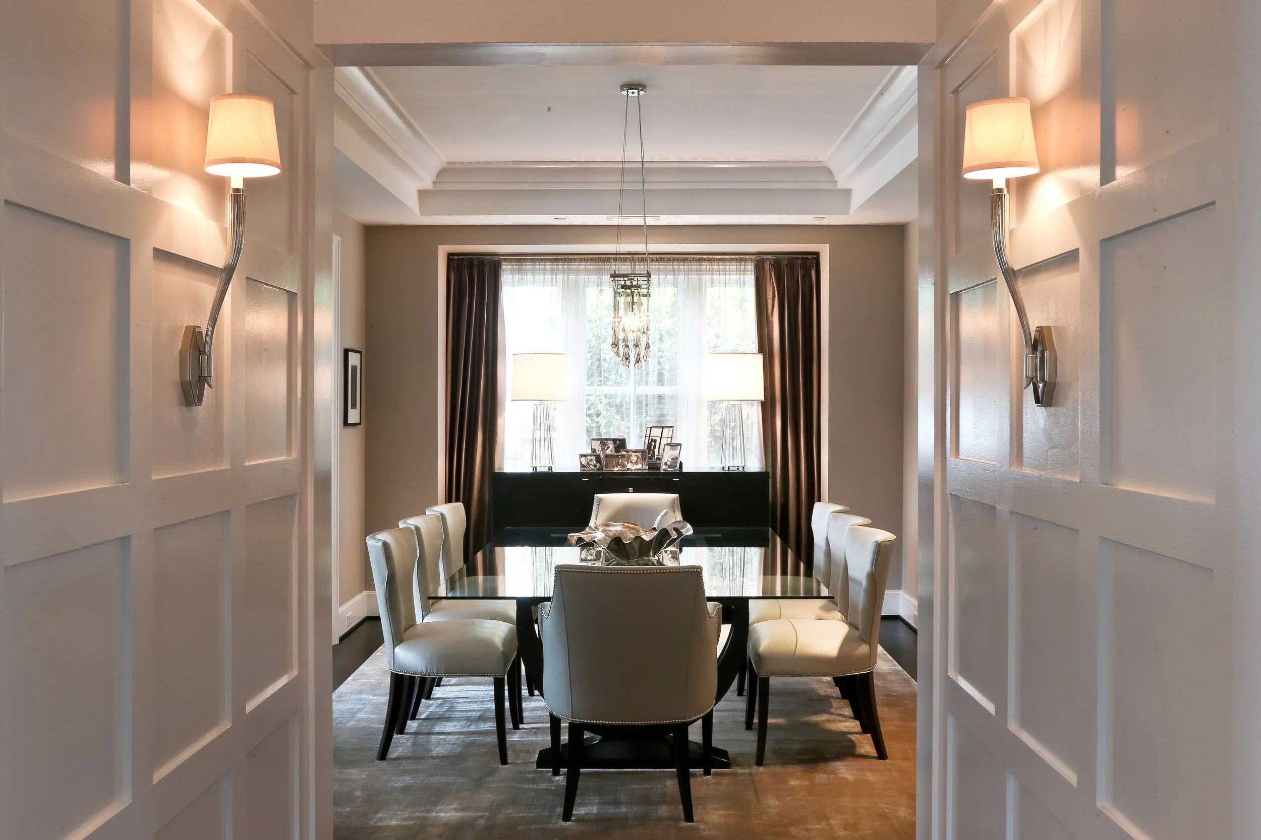 A dining room with a glass-top table surrounded by cream-colored upholstered chairs. A large window with sheer curtains and dark drapes, two table lamps, and a chandelier hang above the table. The room has cream walls and a tray ceiling.