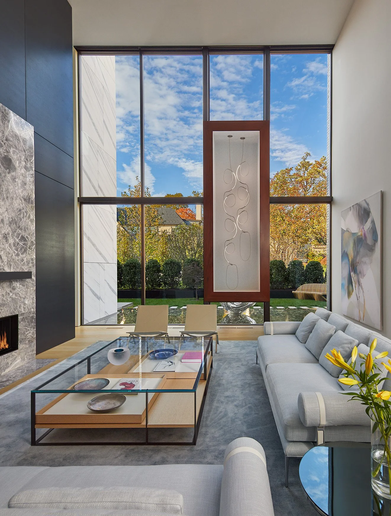 Living room with large floor-to-ceiling window showing a blue sky with clouds and trees outside. Modern furniture includes a light gray sofa, a glass coffee table with decorative plates and books, a beige rug, and an abstract painting on the wall. Th