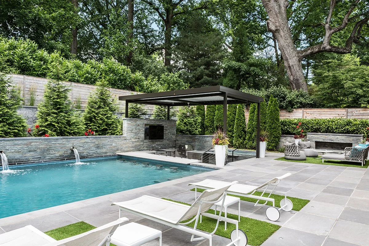 Modern backyard with swimming pool, lounge chairs, outdoor kitchen, potted plants, and a green backdrop of trees and shrubs.