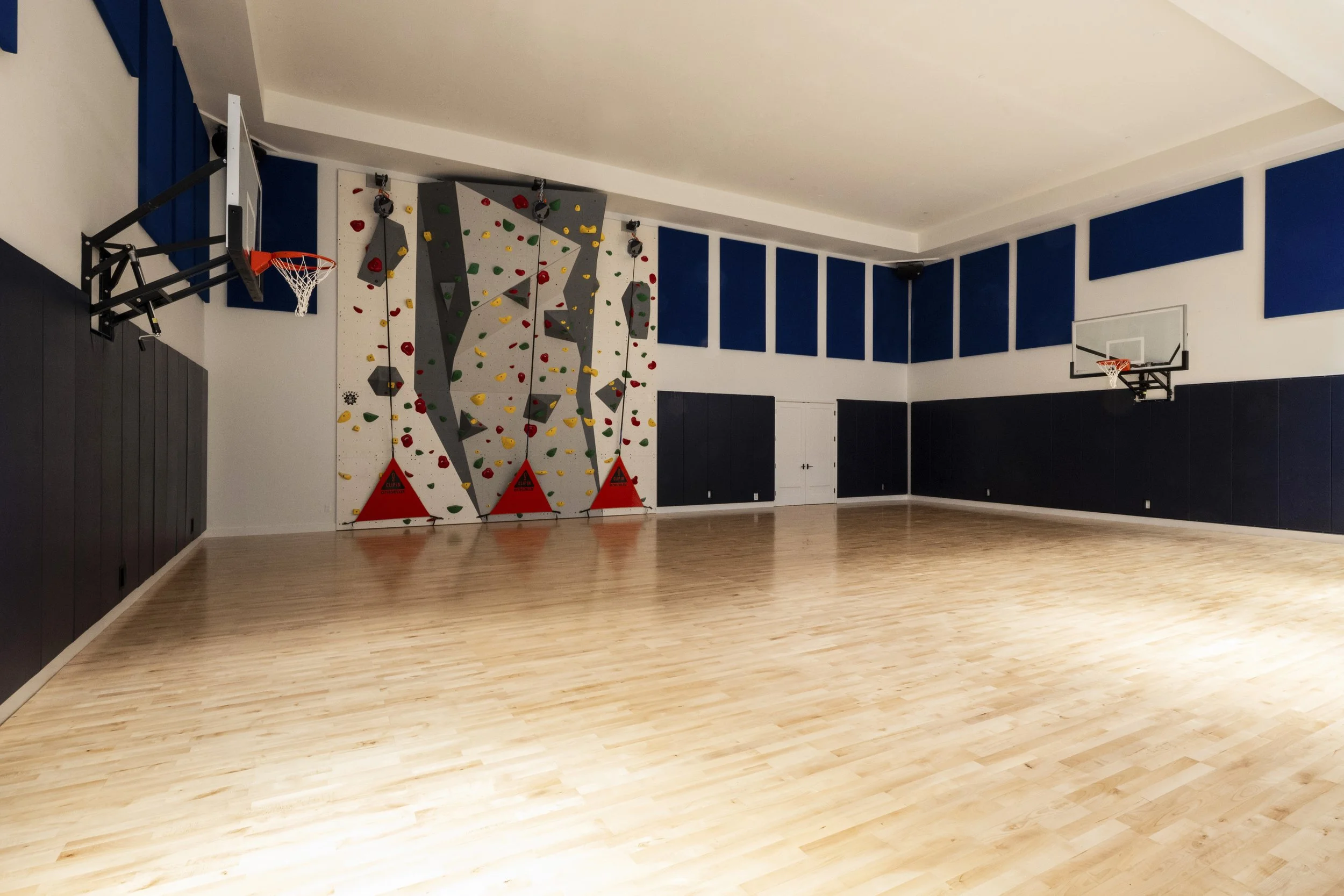Indoor gymnasium with wood flooring, two basketball hoops, and a colorful climbing wall on the left.