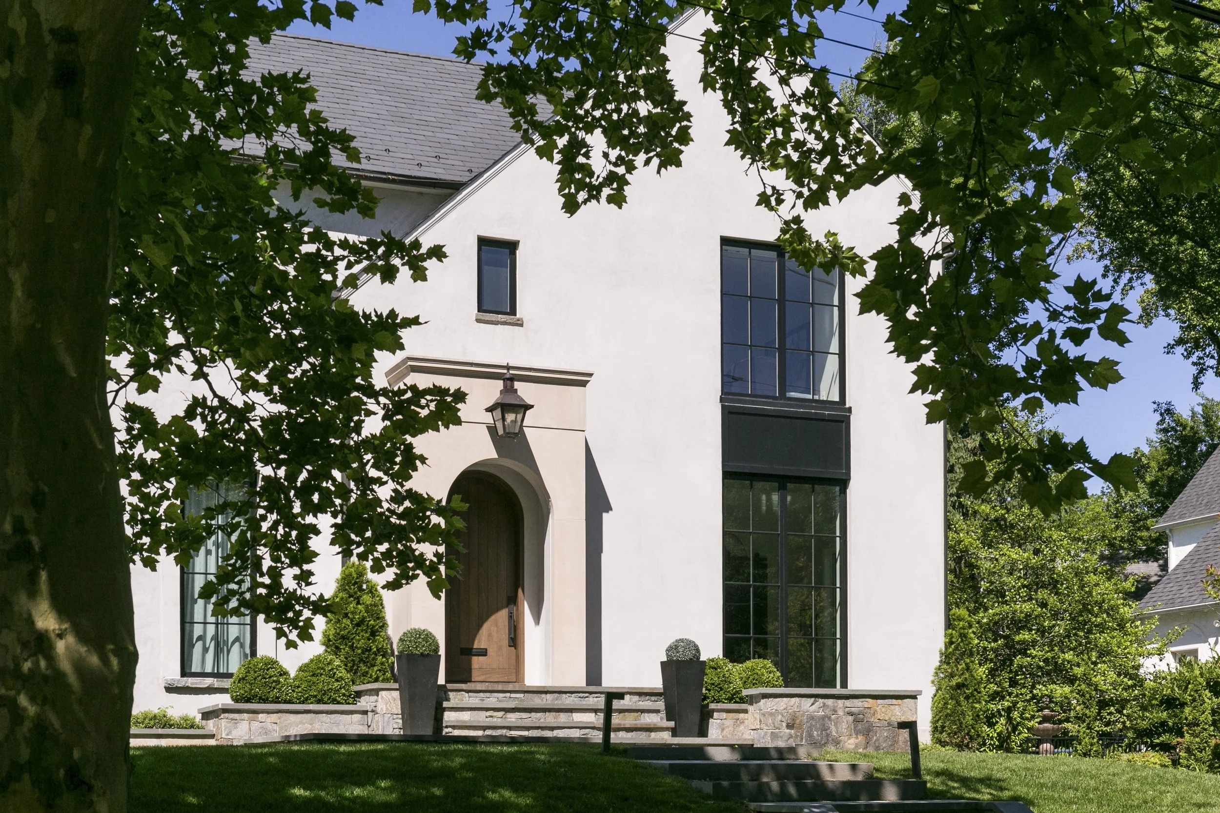White house with large windows, a wooden door, and a stone foundation, seen through green tree branches in a well-manicured yard with potted plants and bushes.