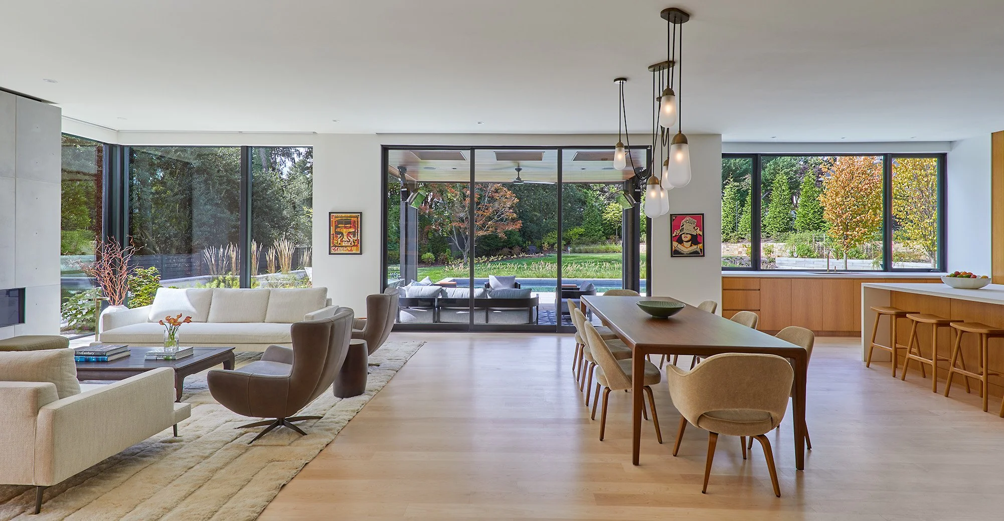 Bright open living room and kitchen with large windows showcasing a lush green yard with trees and outdoor patio, modern furniture, pendant lighting, and minimalistic decor.
