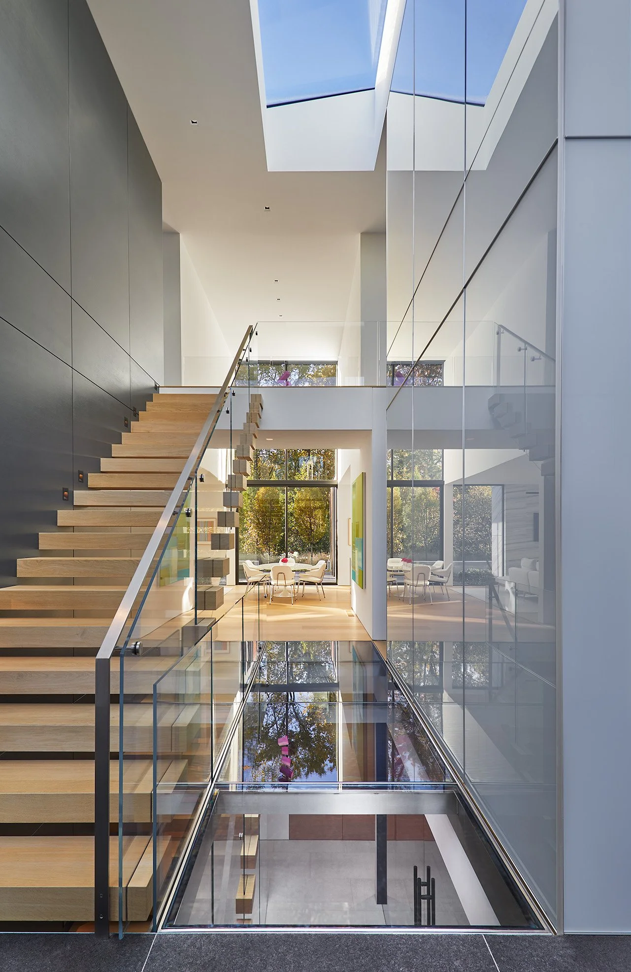 Modern interior of a multi-story building with a glass floor, staircase, and high ceiling with skylights, showing a lobby and seating area with large windows and trees outside.