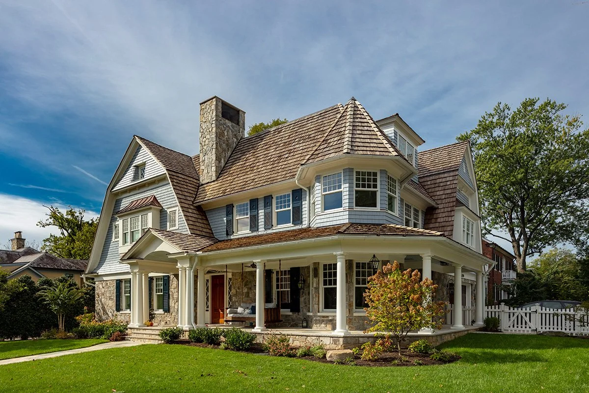 Large, historic-style house with a wraparound porch, multiple gabled roofs, and a stone chimney, surrounded by well-maintained lawn and trees.