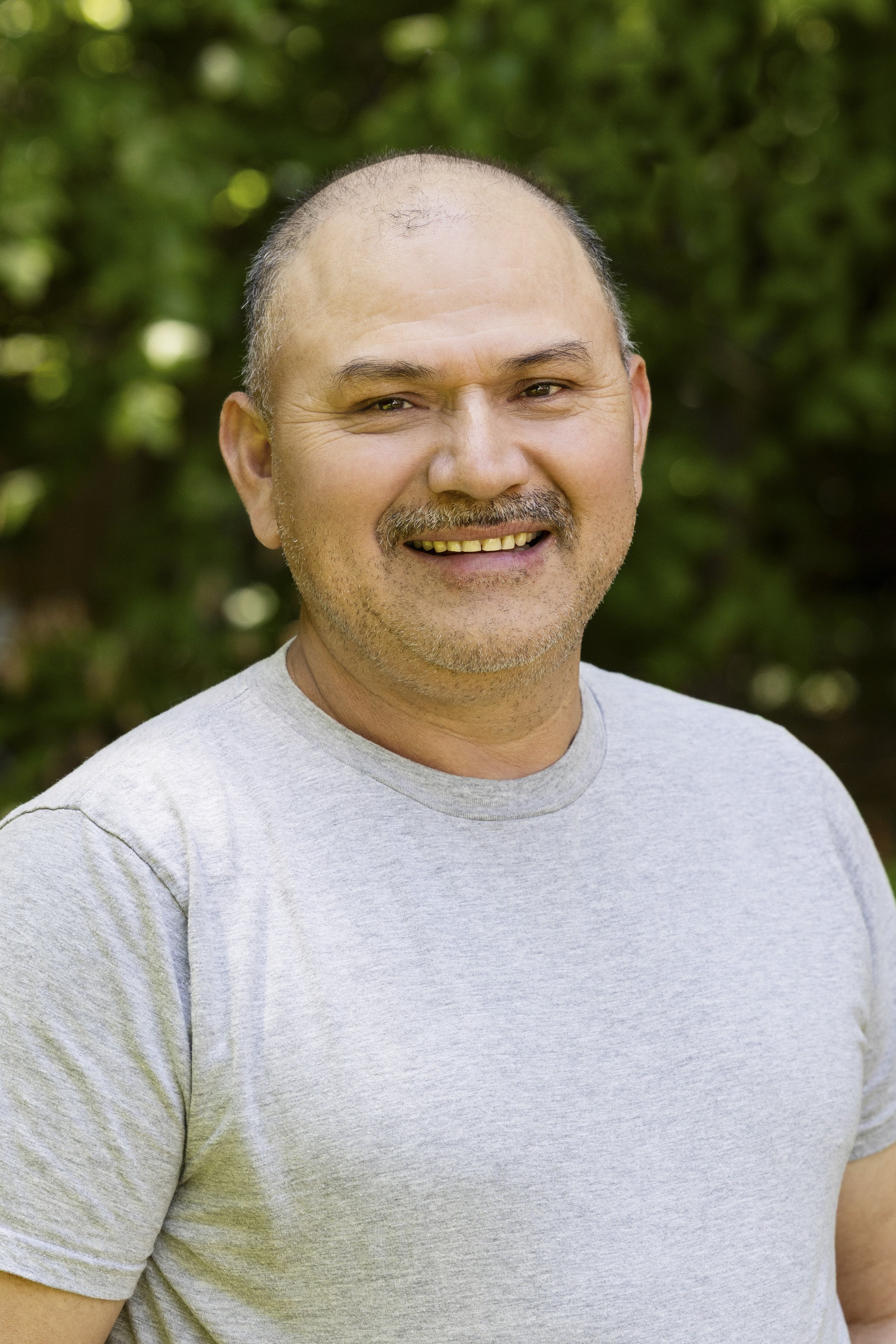 A middle-aged man with a shaved head and a goatee, smiling, wearing a light gray T-shirt, standing outdoors with a blurred green leafy background.