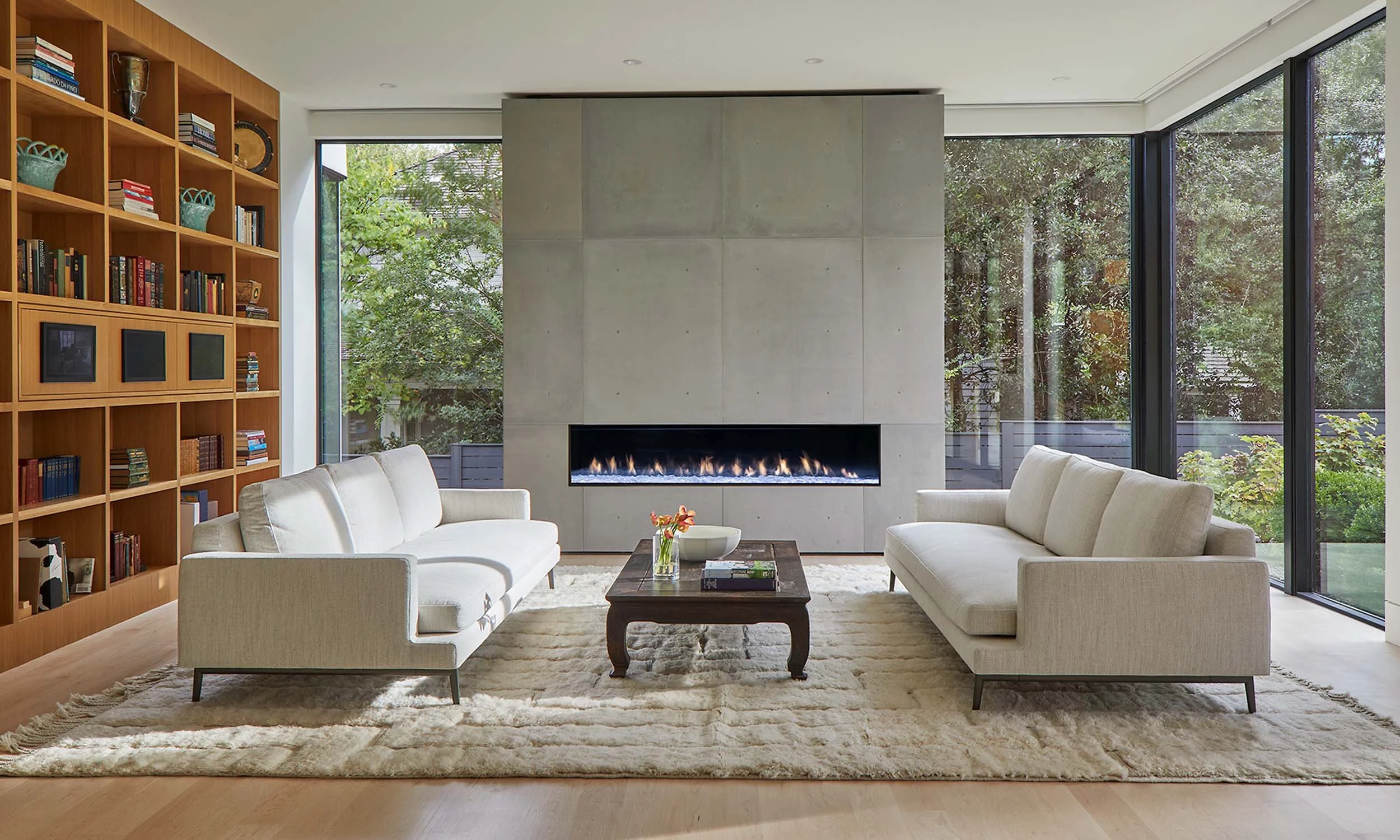 Modern living room with large floor-to-ceiling windows, a fireplace, white sofas, a wooden coffee table, a bookshelf, and a beige area rug.