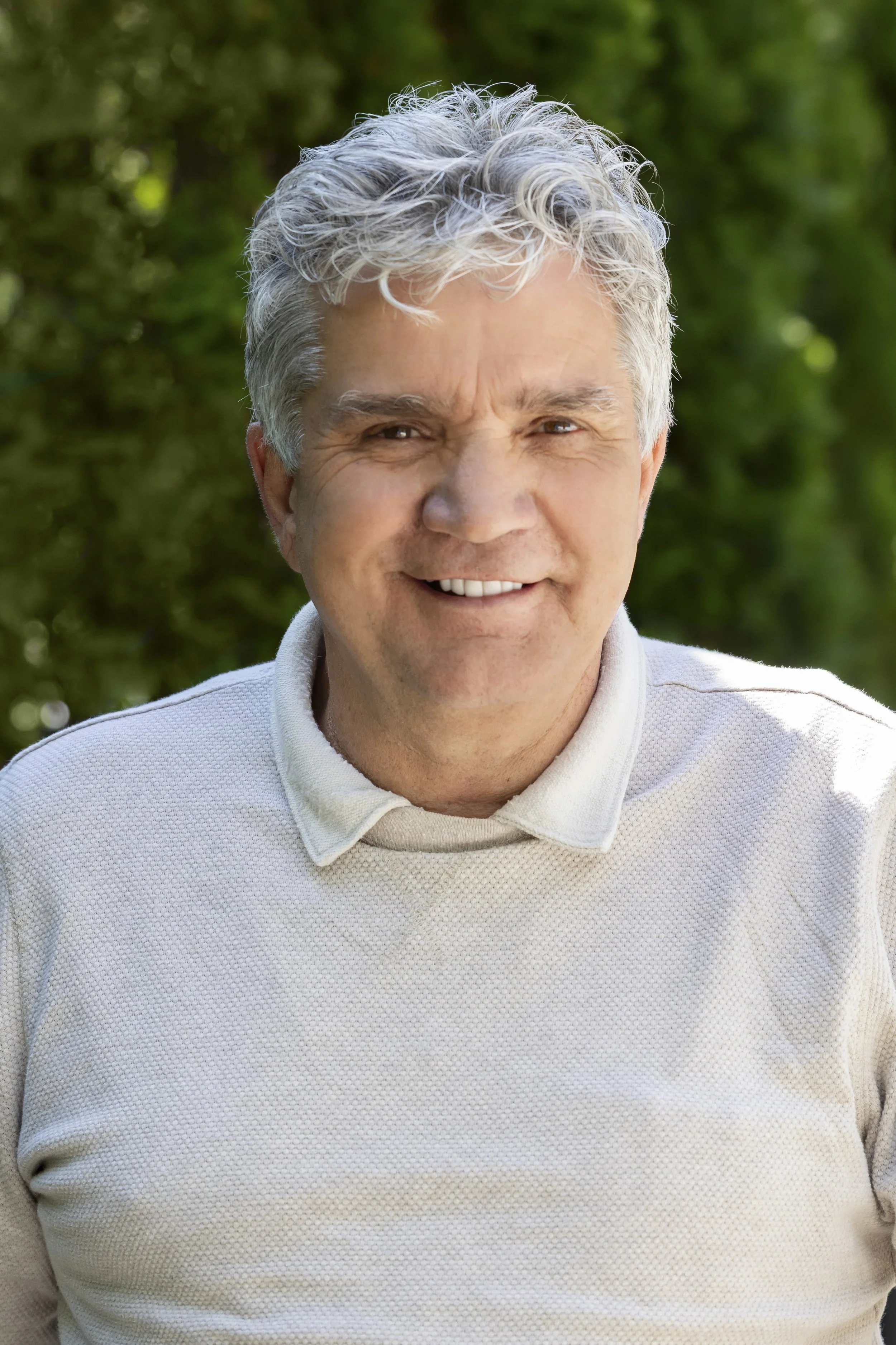 Close-up photo of a smiling senior man with gray, curly hair, wearing a light-colored, textured sweater, outdoors with green blurred foliage in the background.