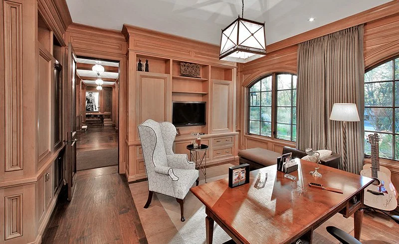 Living room with built-in wood paneling, large windows with curtains, a wooden desk with picture frames and a pen, an upholstered armchair, a floor lamp, and an acoustic guitar. A hallway with pendant lights is visible through the doorway.