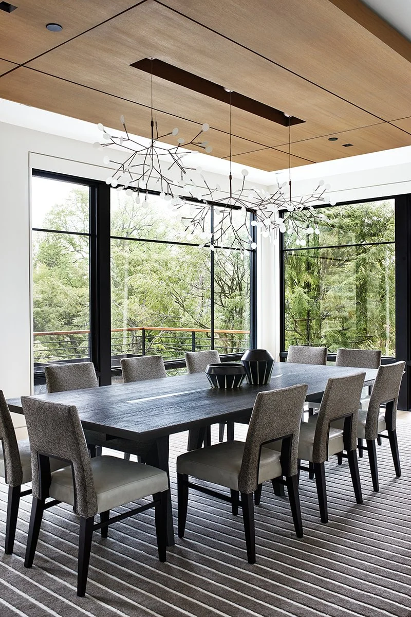 Modern dining room with large windows, a long wooden table, ten upholstered chairs, and a unique pendant light fixture