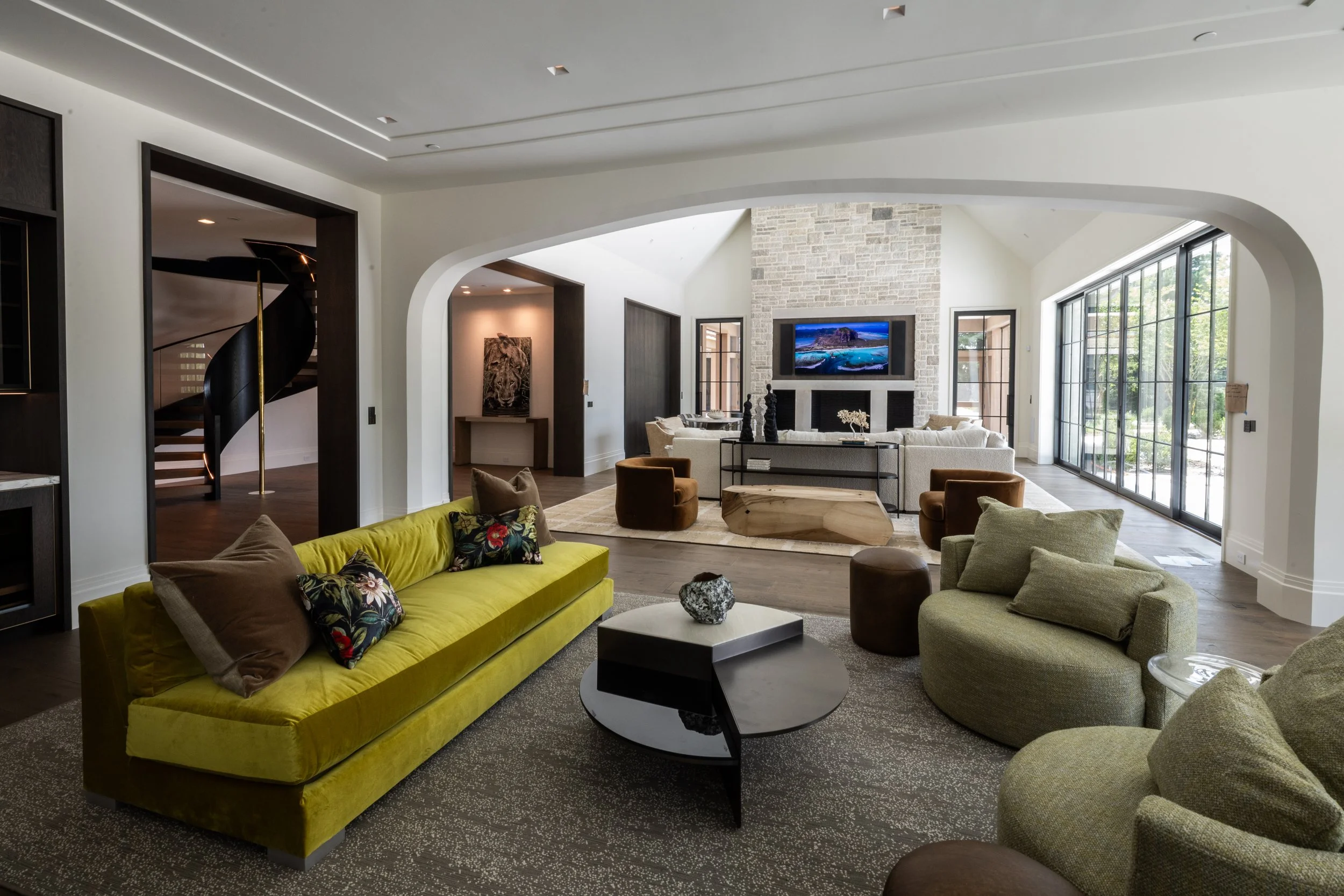 A spacious, modern living room with large windows, a stone fireplace, and contemporary furniture including a yellow velvet sofa, beige and green armchairs, and various decorative items.