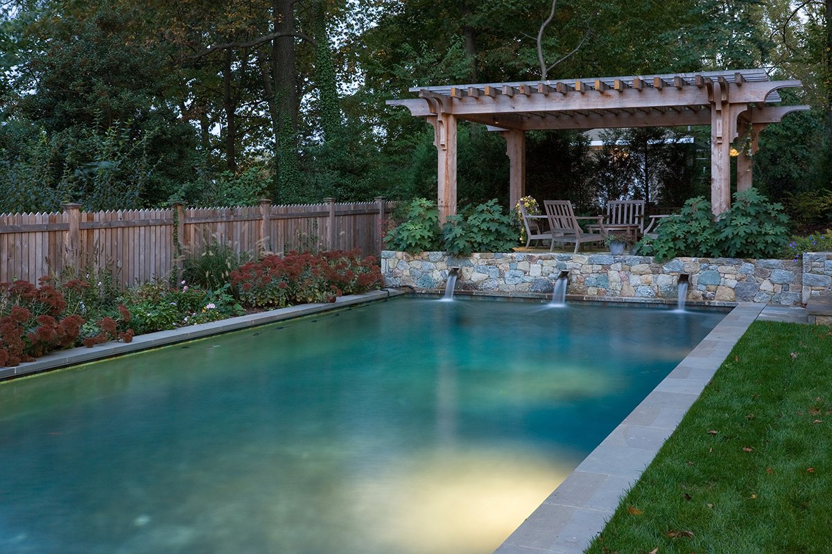 A backyard swimming pool with a garden and a wooden pavilion with benches, surrounded by a wooden fence and trees.