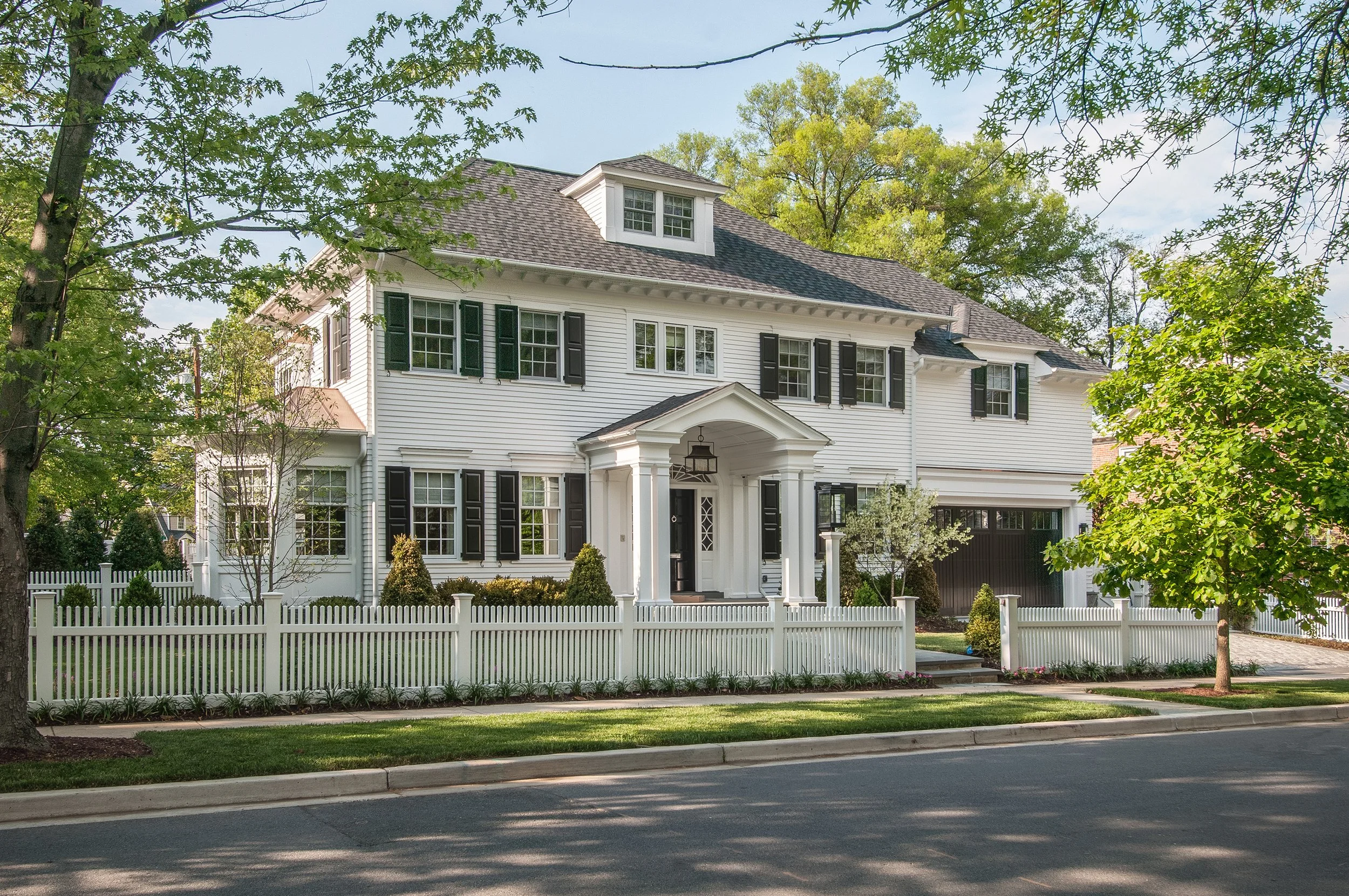 A large white house with black shutters, a covered front porch, and multiple windows, surrounded by a white picket fence, trees, and greenery under a clear sky.