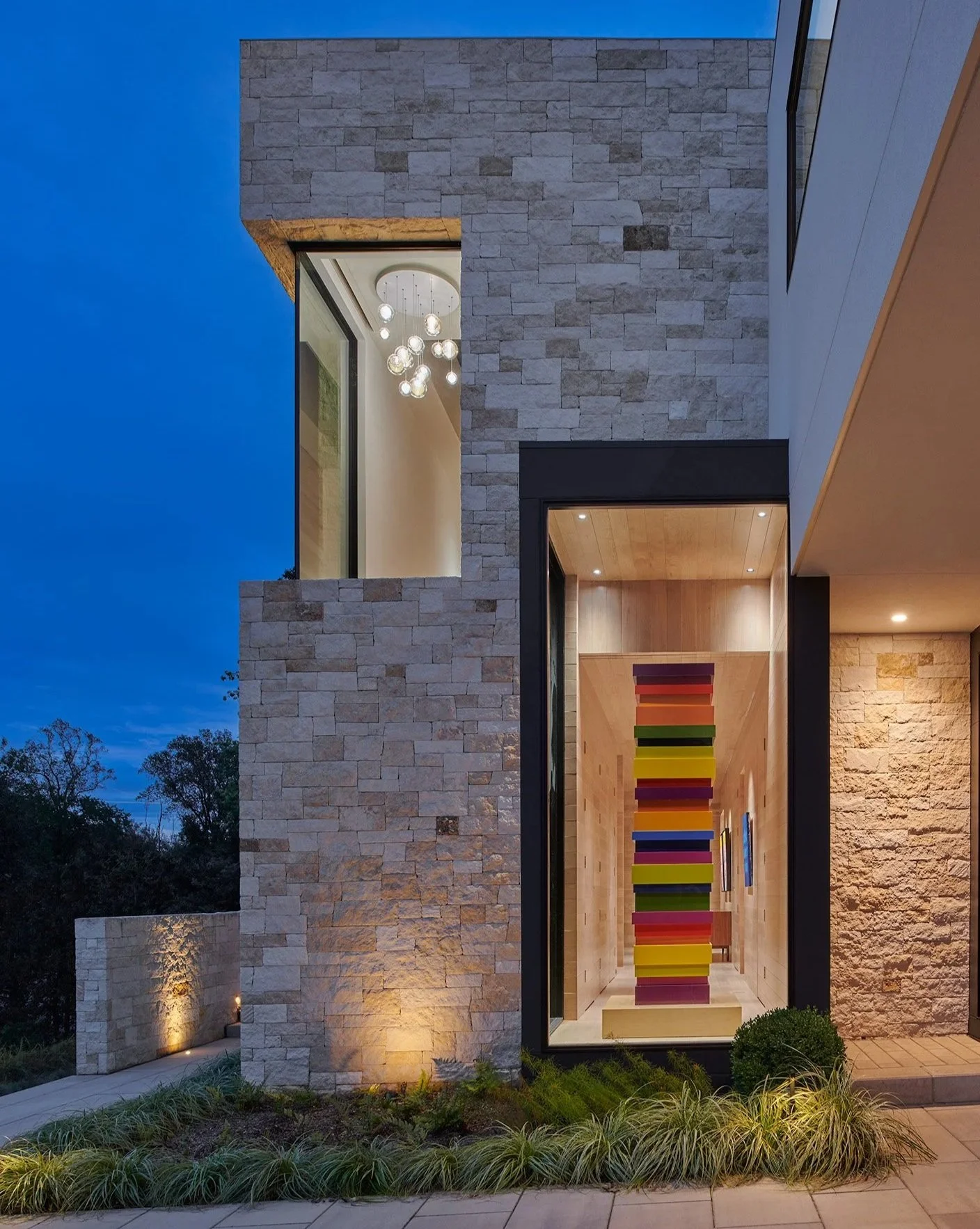 Exterior of a modern building with stone walls and large window. Inside, a colorful abstract sculpture with stacked rectangular shapes extends vertically. The scene is illuminated by outdoor lighting at dusk, with trees and plants in the surrounding 