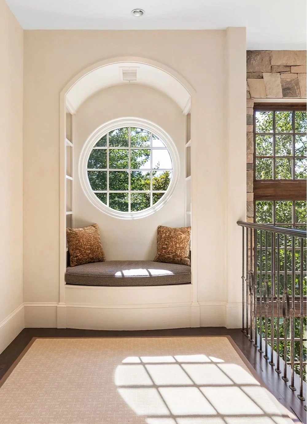 Sunlit built-in window seat with cushions and round window with grid pattern, overlooking greenery outside, in a bright indoor space.
