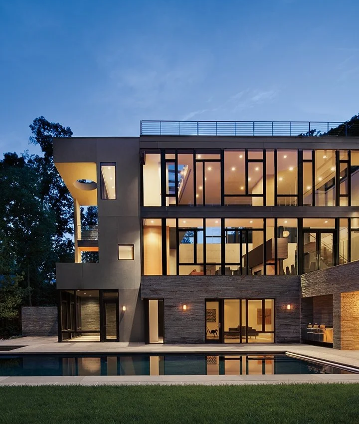 Modern multi-story house with large glass windows, a pool in the backyard, and trees surrounding the property at dusk.