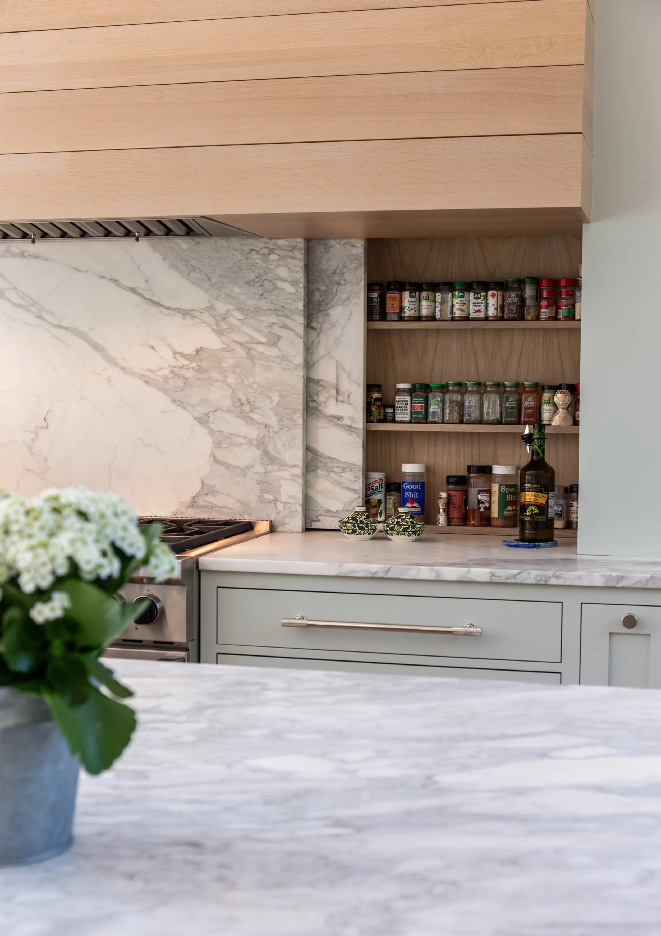 Close-up of a kitchen with a white marble countertop, a gray cabinet drawer, a and a potted green plant with white flowers in the foreground. The background shows a wooden shelf with spice jars and a bottle of olive oil, a marble backsplash, and a bu