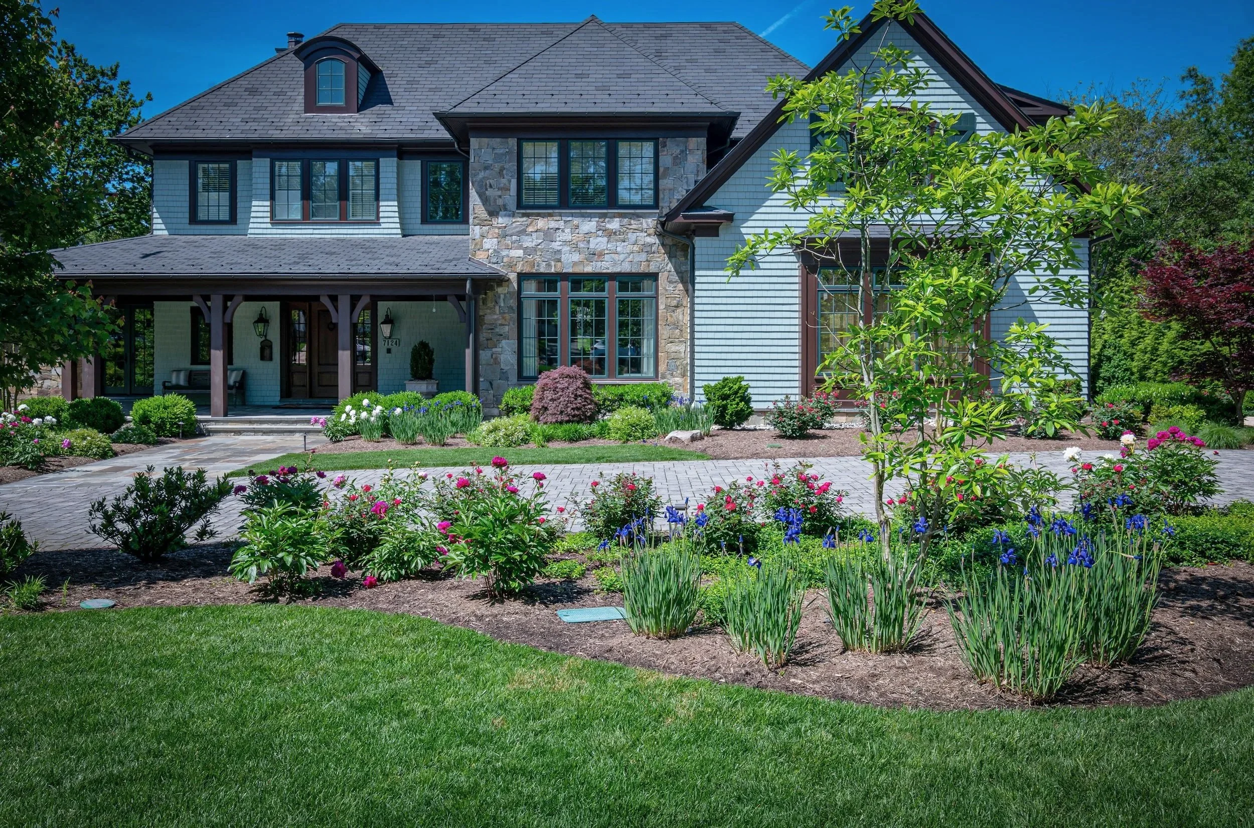 A modern two-story house with a stone and light blue siding exterior, black framed windows, and a dark gray roof, surrounded by a landscaped garden with colorful flowers, bushes, and green trees under a bright blue sky.
