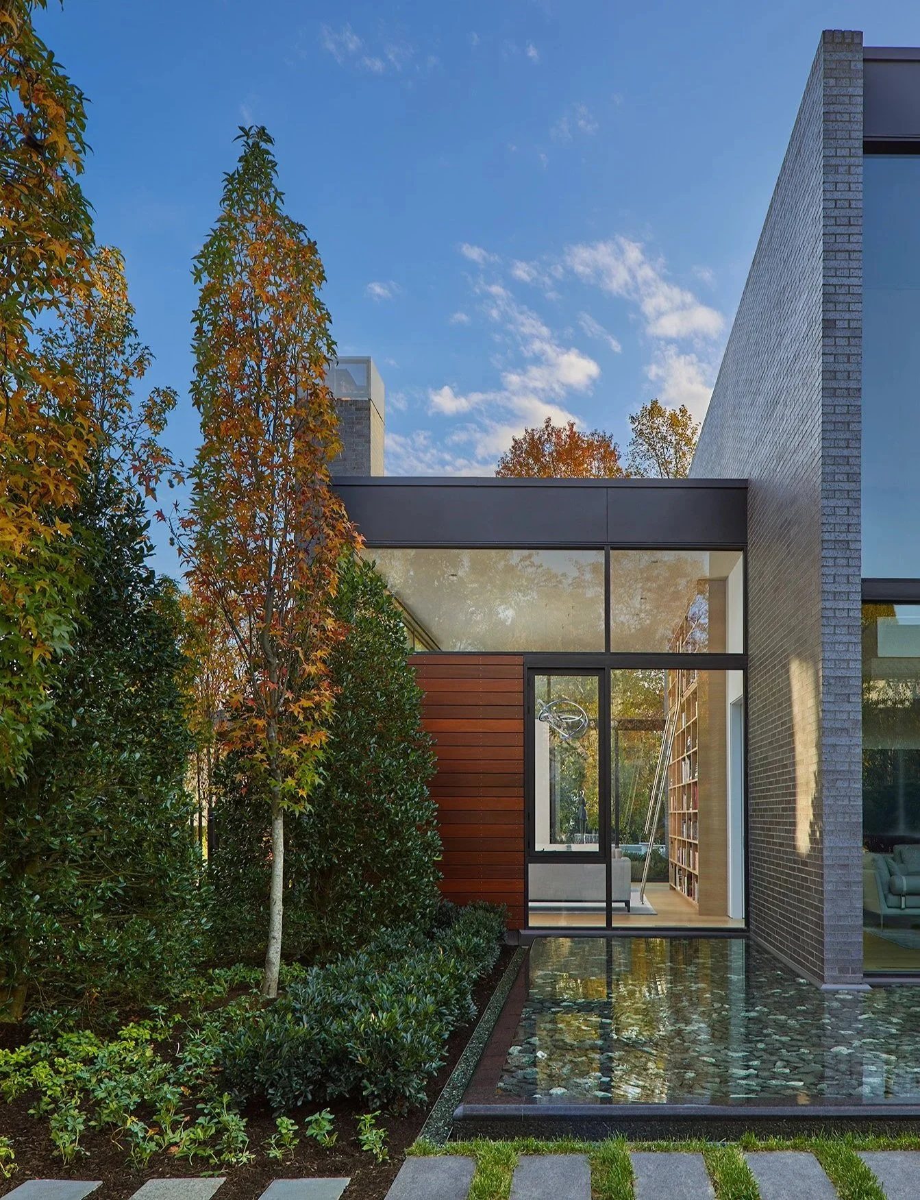Modern house with large glass windows, surrounded by trees and shrubs, featuring a pond with stones in the foreground and a pathway leading to the house.