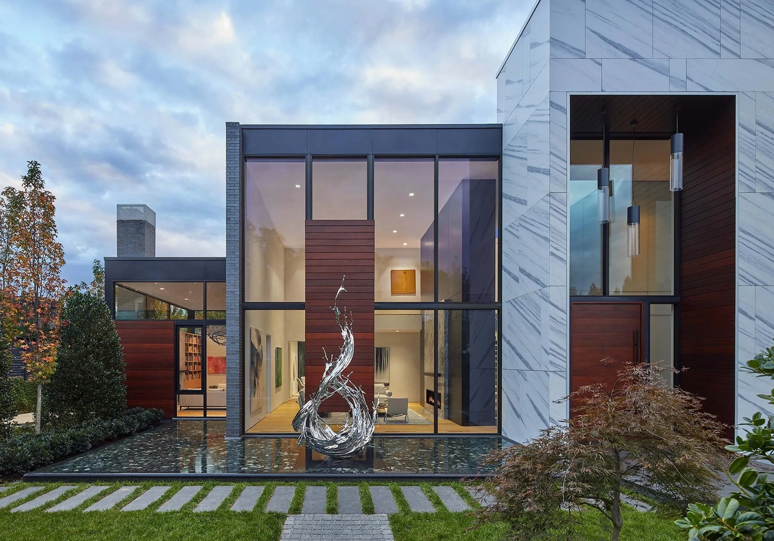 Modern house with large glass windows, marble siding, and a water fountain sculpture in the front yard, surrounded by trees and a lawn.