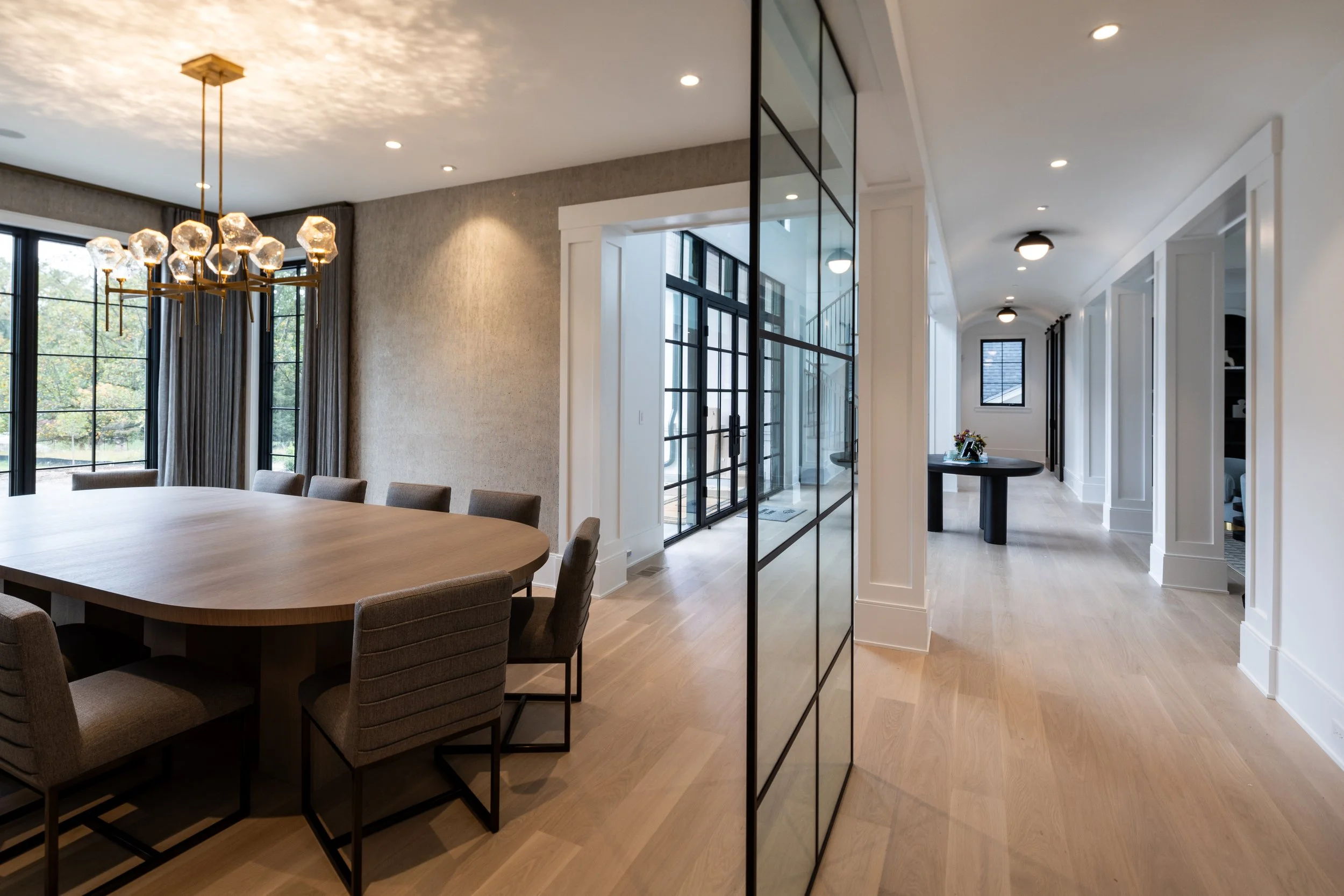 A modern dining room with a wood table and upholstered chairs. Large windows with black frames and gray curtains offer a view of trees outside. A multi-globe chandelier hangs above the table. A glass door with black frames leads to an outdoor area, a