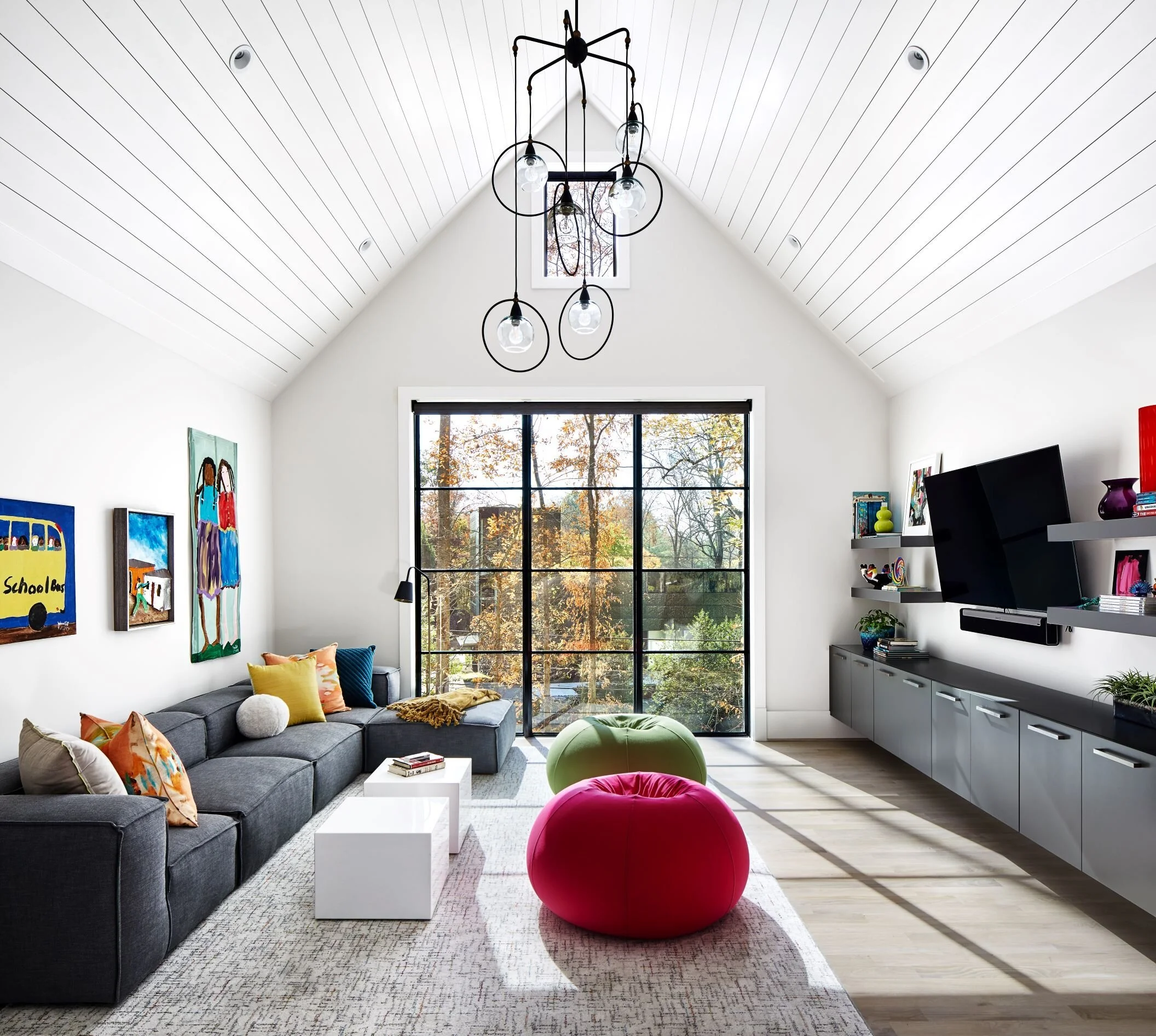 Bright living room with large window, gray sectional sofa with colorful pillows, modern chandelier, artwork on white walls, white side tables, and a red pouf.
