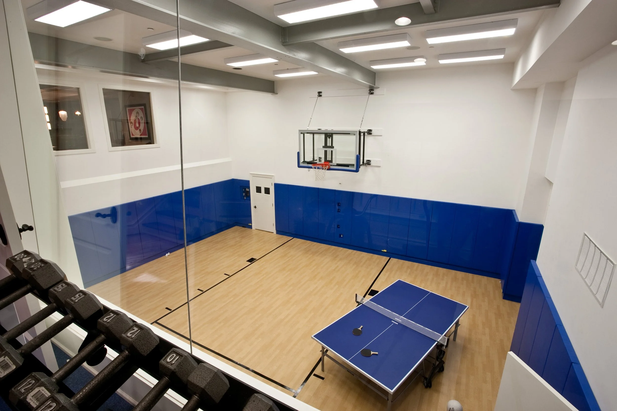 Private indoor basketball court with blue padded walls, a mounted basketball hoop, a ping pong table, and a row of dumbbells visible through a glass wall.