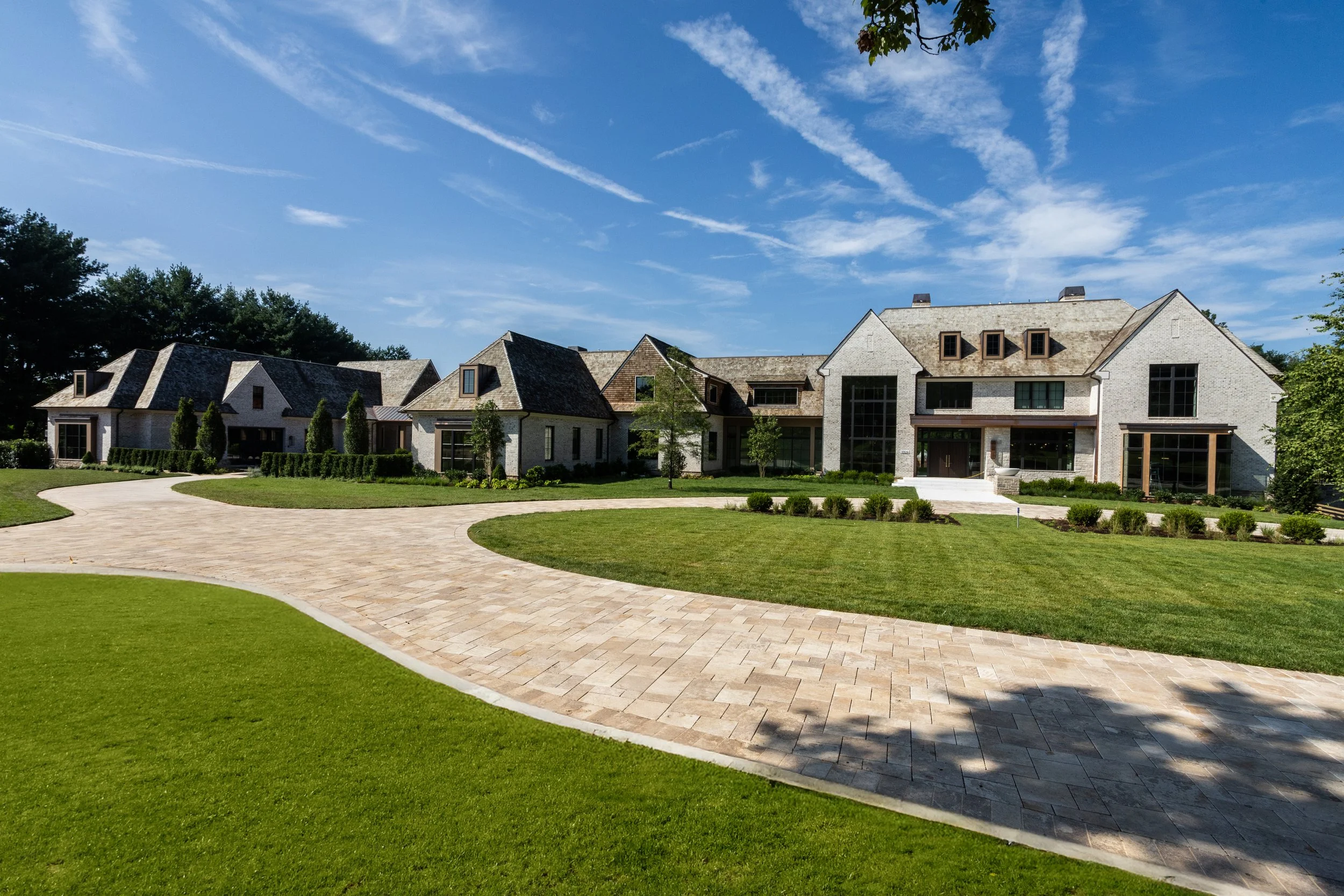 A large modern house with gray brick and shingles, multiple gabled roofs, large windows, and a well-maintained lawn with paved walkway, under a blue sky with scattered clouds.