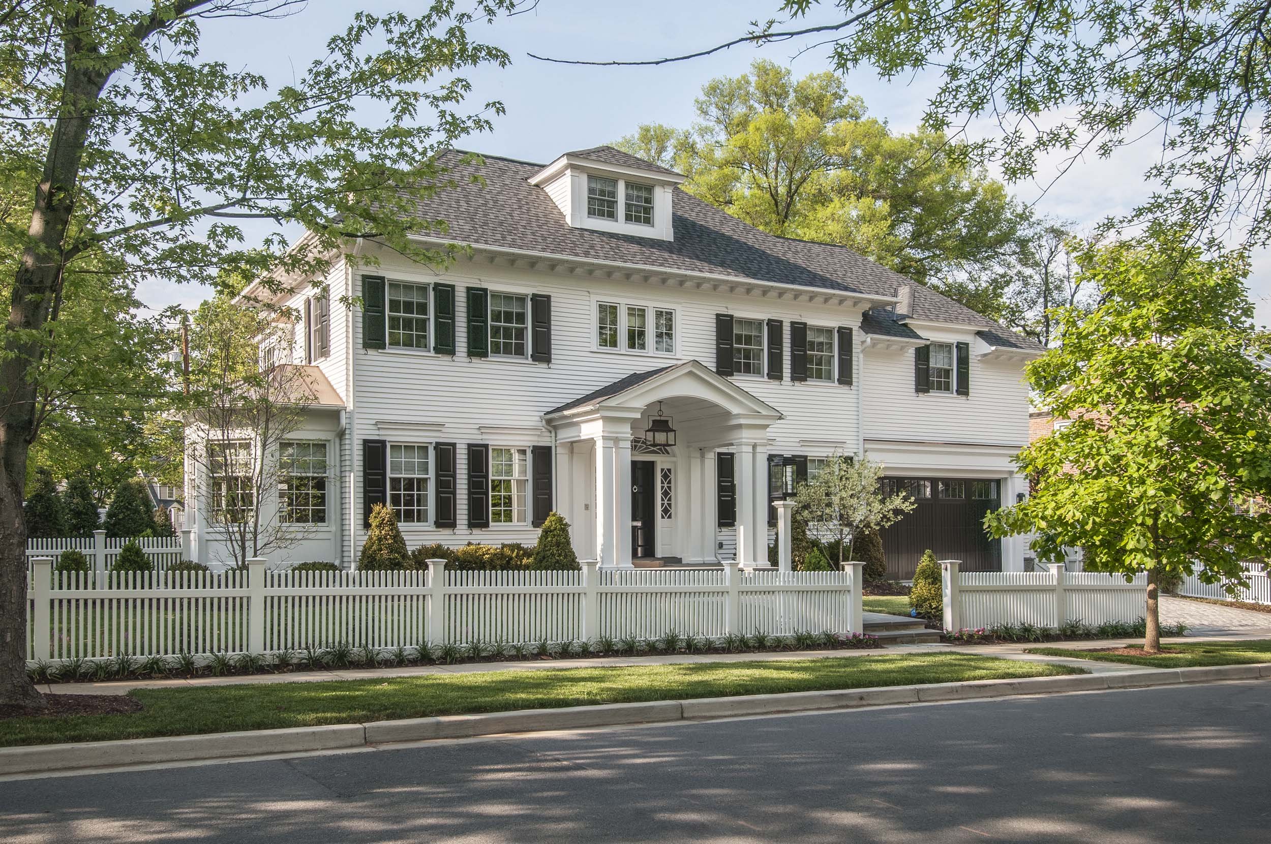 A large white house with black shutters, a front porch with white columns, a white picket fence, and surrounded by green trees and lawn, with a street in front.