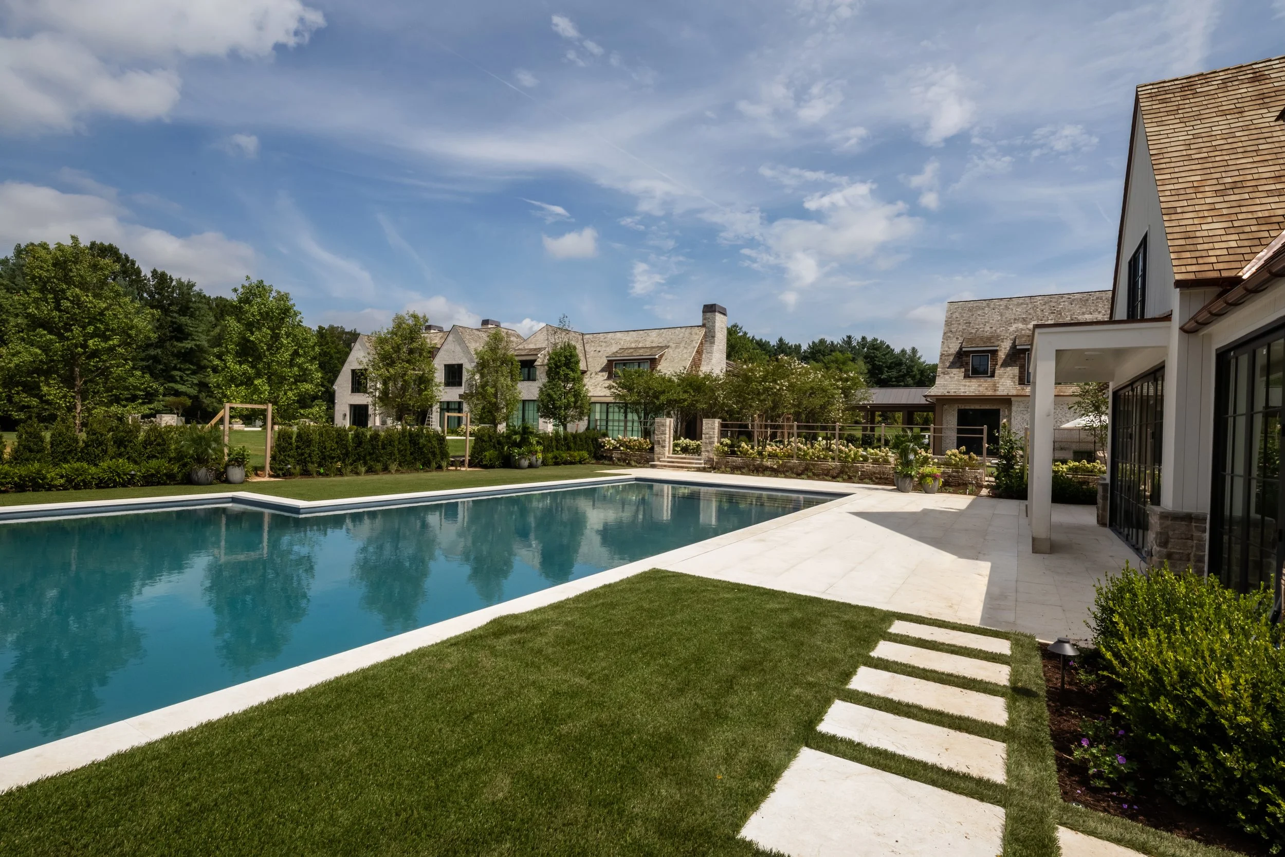 A backyard with a swimming pool, lush green grass, and a row of trees behind a modern residential house with multiple gables and large windows. The sky is partly cloudy with blue and white clouds.
