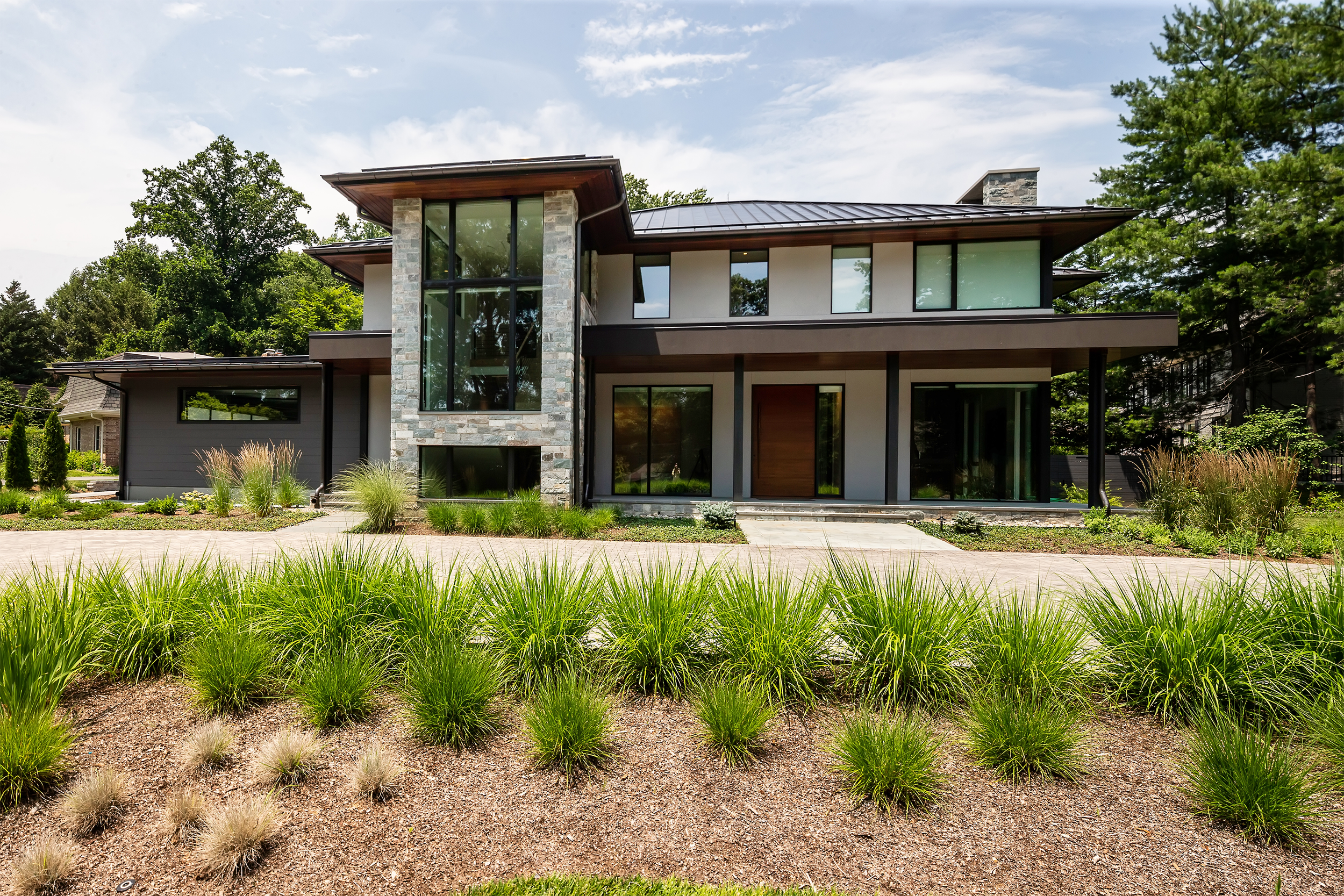 Modern two-story house with large glass windows, stone and stucco exterior, surrounded by well-maintained landscaping and tall trees.