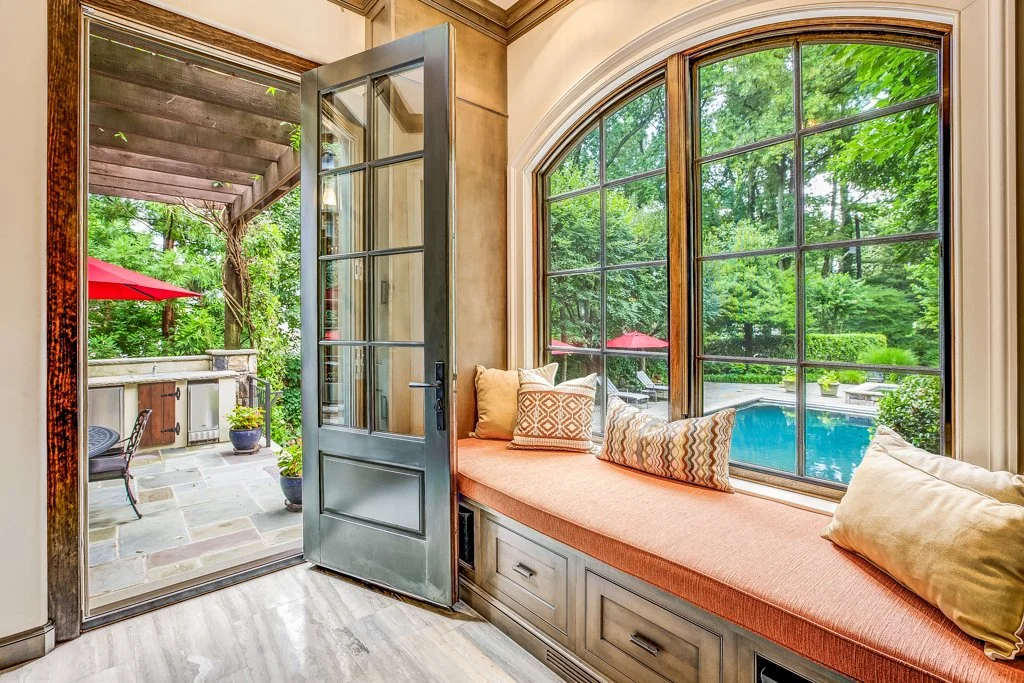 Indoor window seat with cushions overlooking a backyard pool and patio with red umbrellas