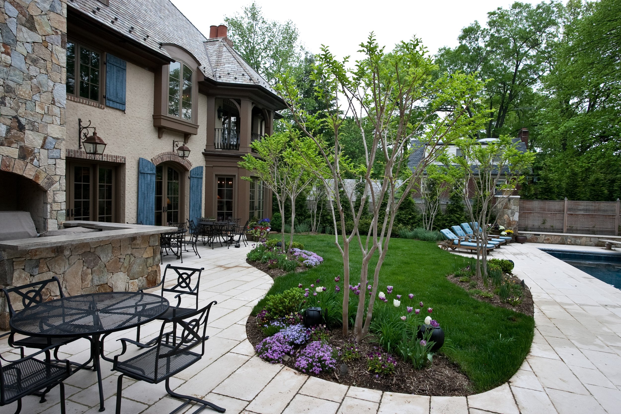 A backyard patio with black metal tables and chairs, a garden with colorful flowers, a swimming pool, and a large house with stone and stucco exterior, blue shutters, and multiple windows, surrounded by lush green trees.