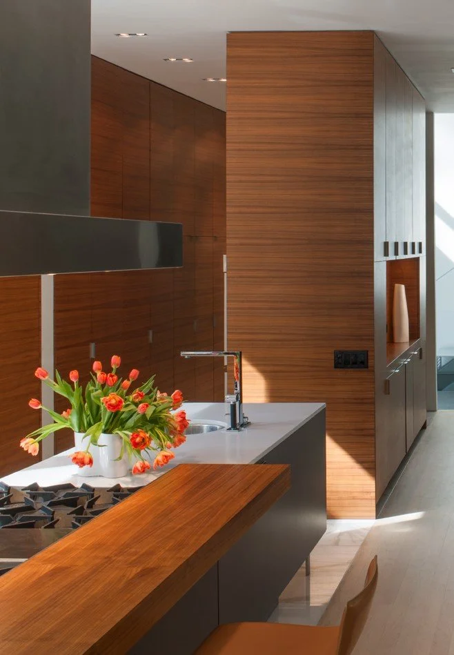 Modern kitchen with wooden cabinets, a white countertop, a gas cooktop, a flower vase with orange flowers, and a chair in the foreground.