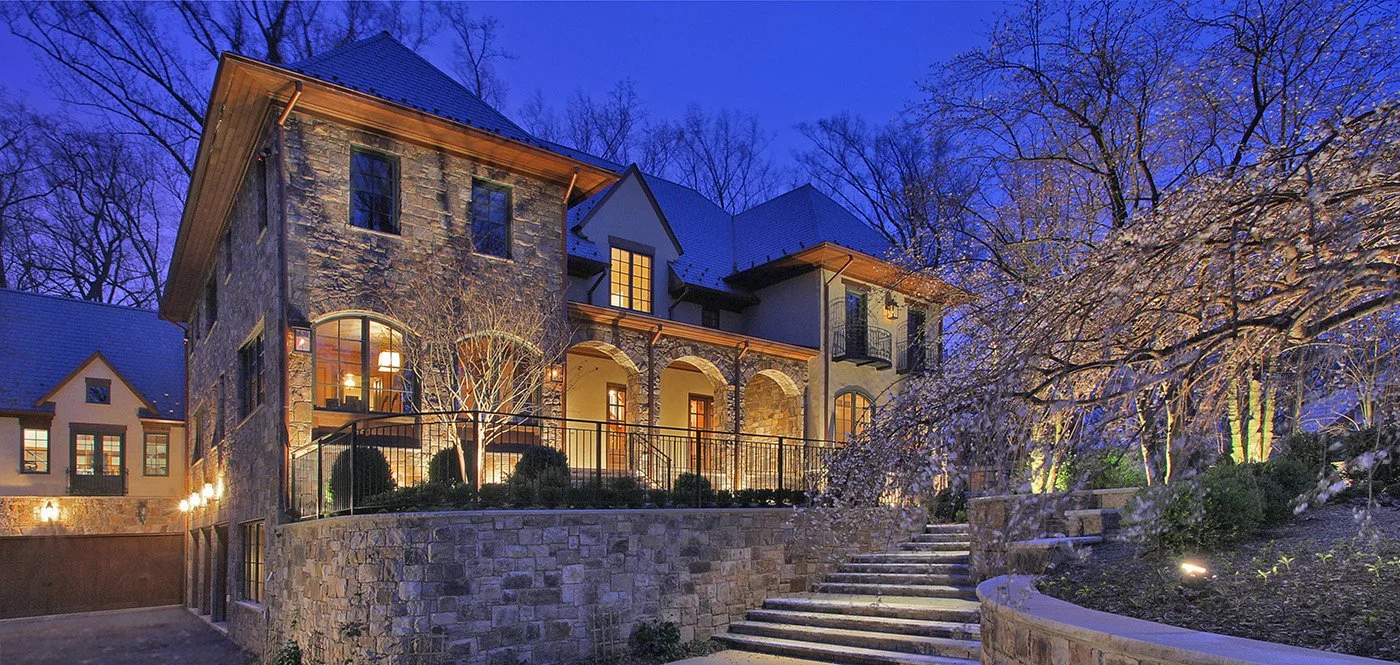 A large, multi-story stone house illuminated at dusk, with trees and landscaped garden in front, stone stairs leading up to the entrance, and a blue sky in the background.