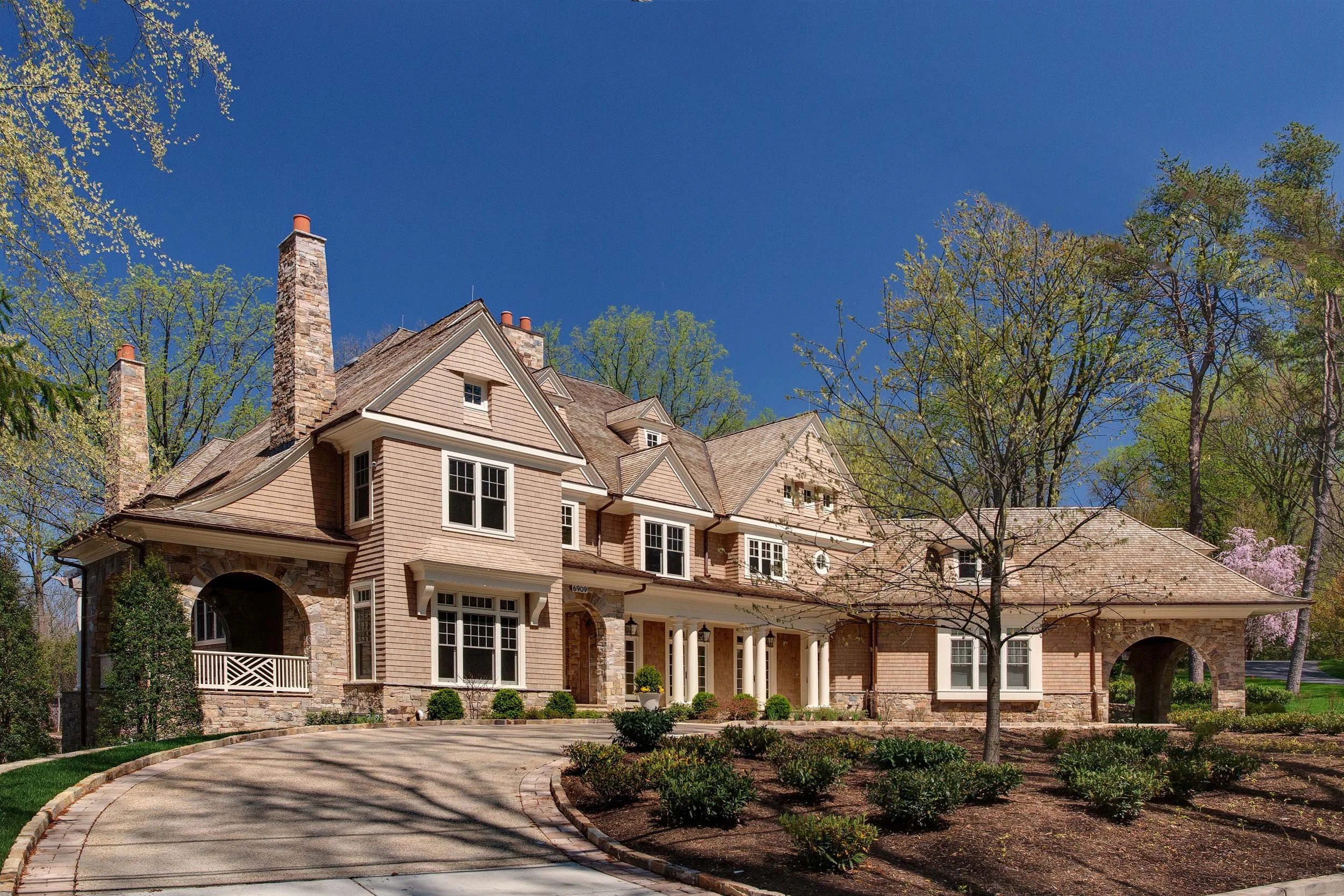 Large house with multiple gables and stone chimney, surrounded by trees and a curved driveway under a clear blue sky.