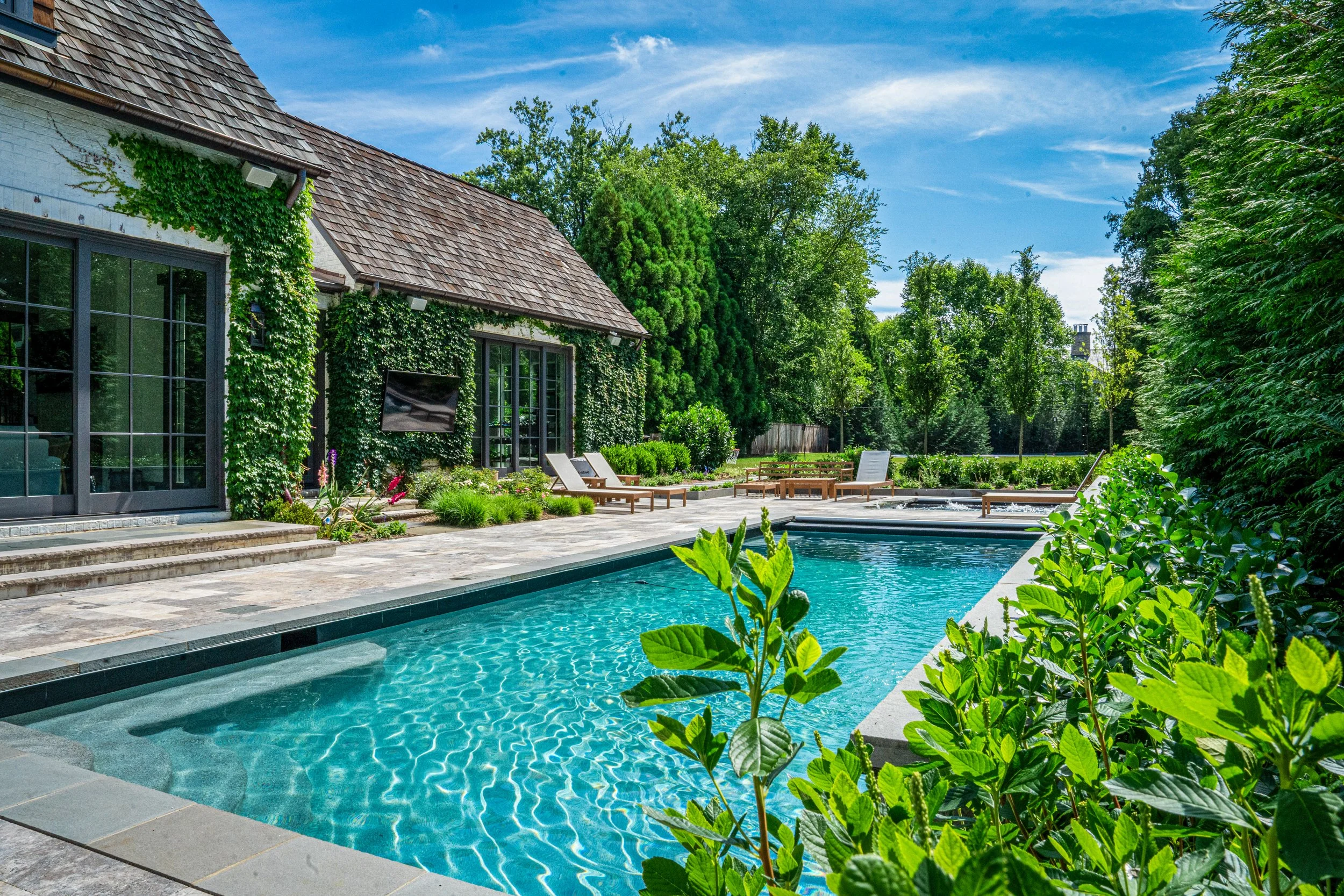 A backyard with a swimming pool, surrounded by green plants and trees, and a house with large windows and a sloped roof covered with shingles.