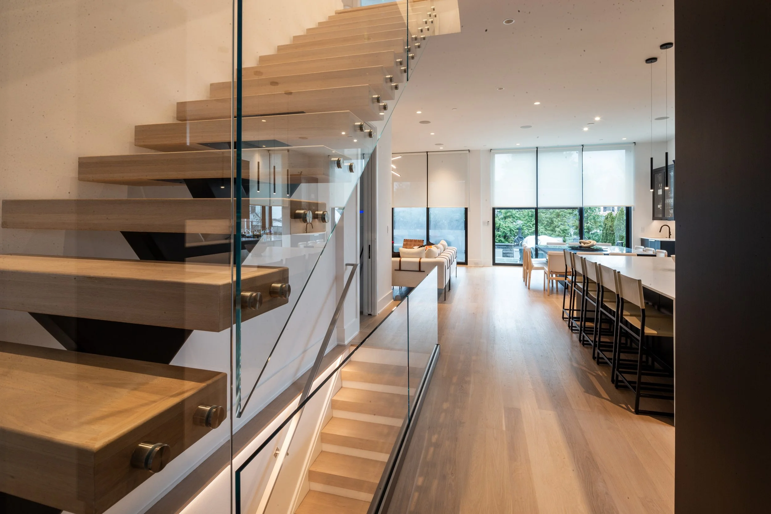 Interior view of a modern, open-plan living space with wooden flooring, large windows with blinds, a staircase with glass railing, and a dining area with chairs.