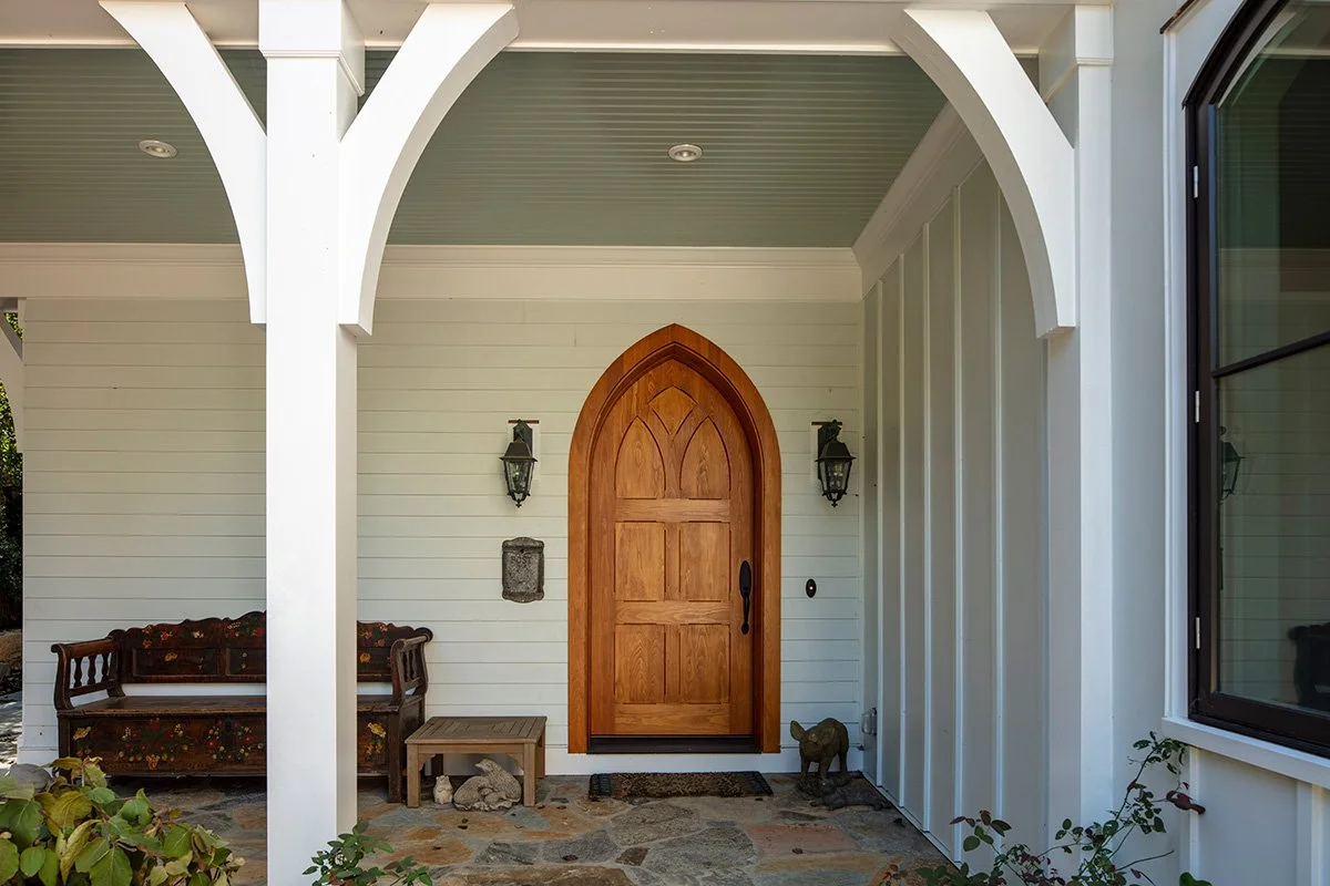 Front porch with a wooden arched door, two wall-mounted lanterns, a porch bench with floral upholstery, a small wooden table, and decorative stones. There's a dog sculpture near the door and a window on the right.