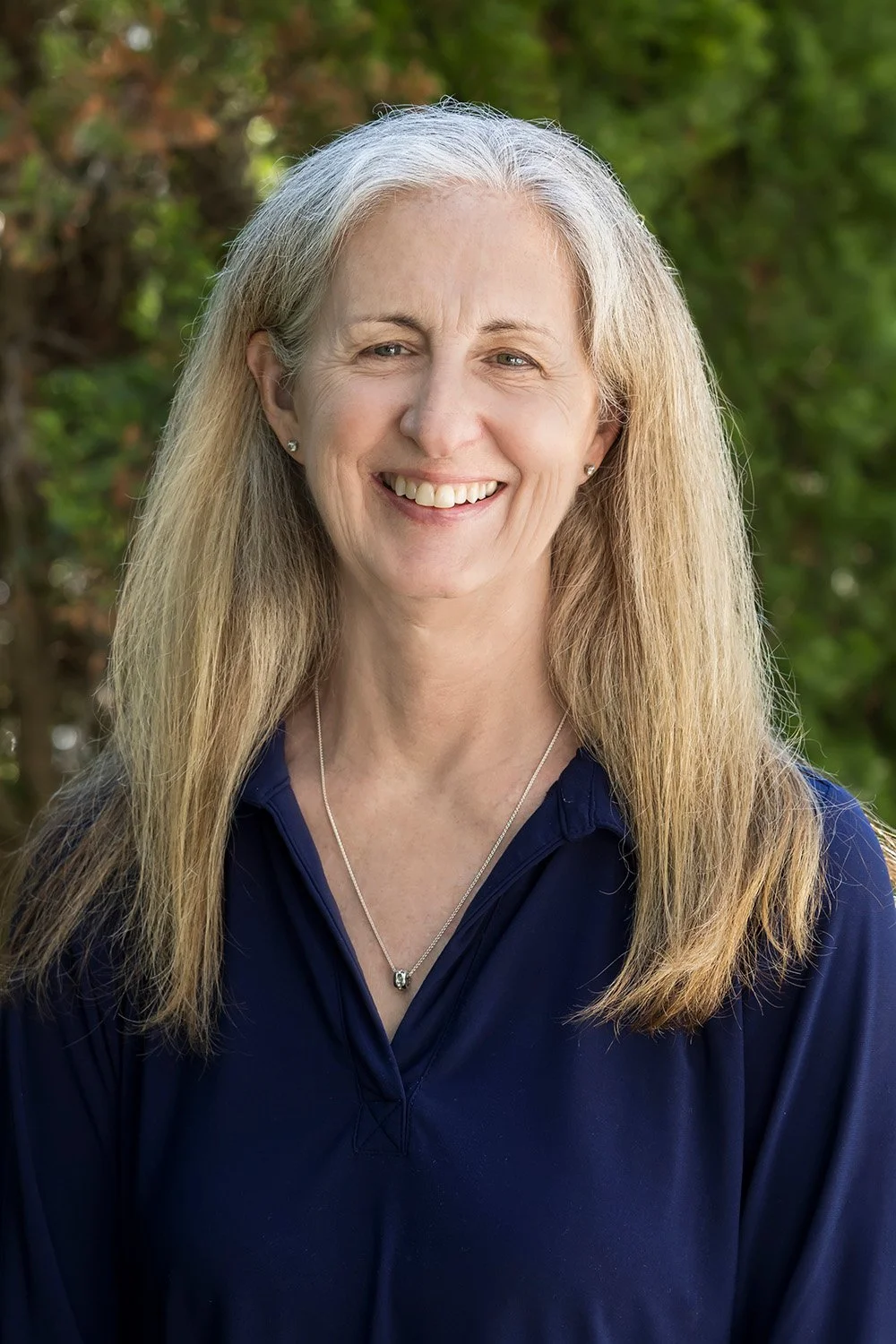 Close-up of a smiling middle-aged woman with long blonde hair, wearing a navy blue top and a silver necklace, outdoors with greenery in the background.
