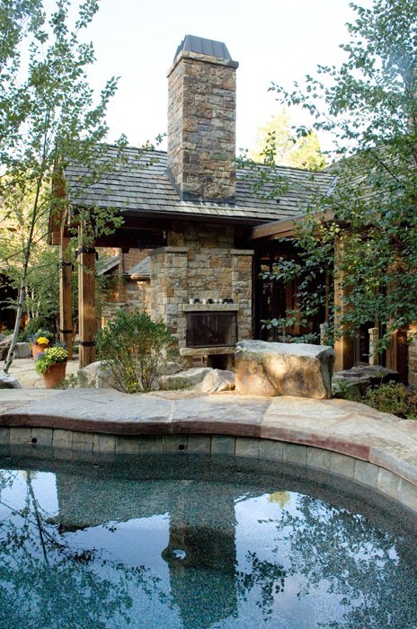 Outdoor stone fireplace beside a pool with trees and patio furniture around.