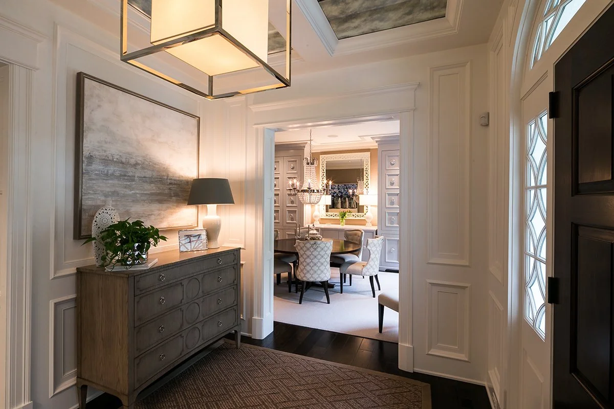 Entryway with white walls, a gray wooden console table with a lamp, decorative objects, and a large abstract painting, leading into a dining area with a round table and upholstered chairs.