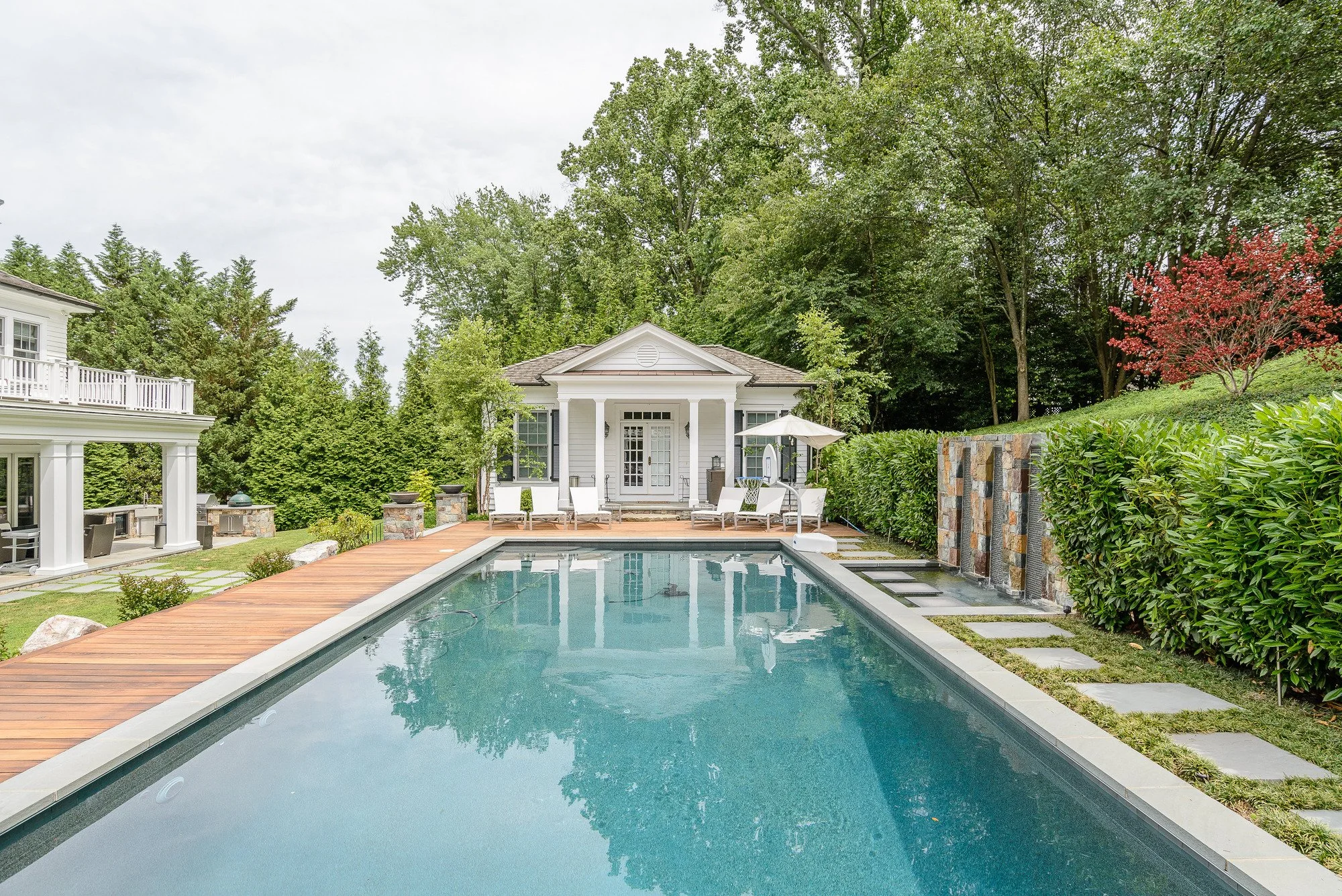 Luxurious backyard retreat with a swimming pool, white house with porch, patio furniture, umbrella, surrounded by trees, bushes, and a stone wall.
