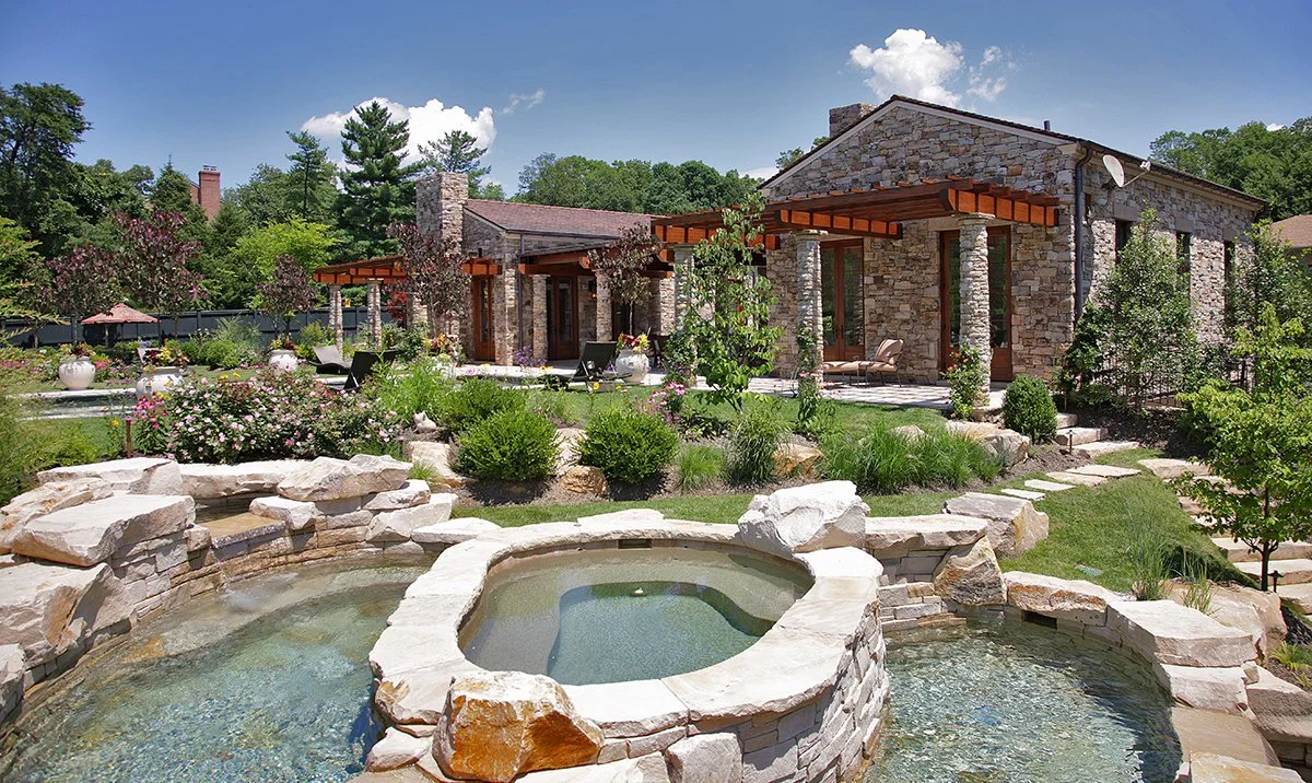 Stone house with covered patio, lawn, trees, and a stone water feature in front.
