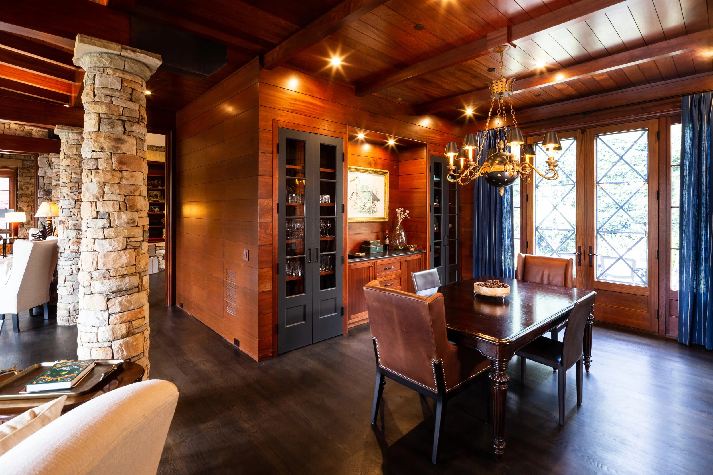 Interior of a wooden house with a dining area, large windows with blue curtains, a chandelier, and a stone pillar.
