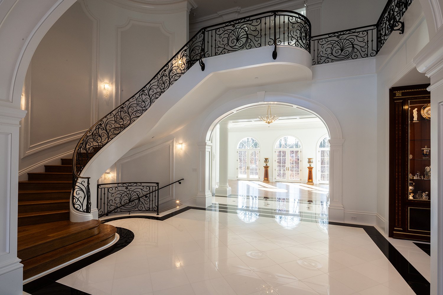 Elegant foyer of a mansion with a grand staircase featuring wrought iron railings, white walls with decorative molding, a glossy white tiled floor with black accents, and a sunlit sitting area with large arched windows and gold decorative vases.