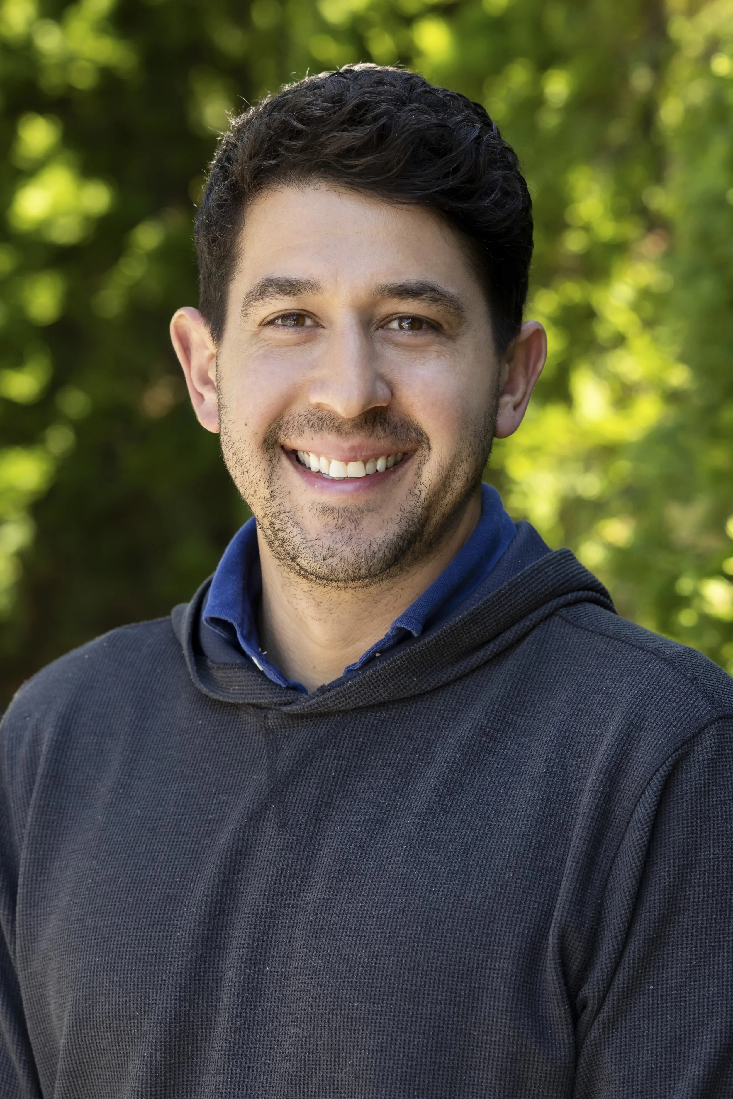 A man smiling outdoors in front of a green, blurred background.